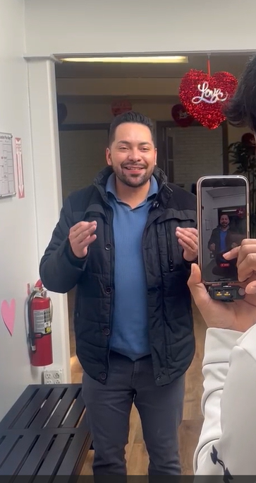 Hombre sonriendo y posando para una foto en un lugar con decoración de Día de San Valentín, con un corazón rojo que dice "Love" y un tacón de tacón.