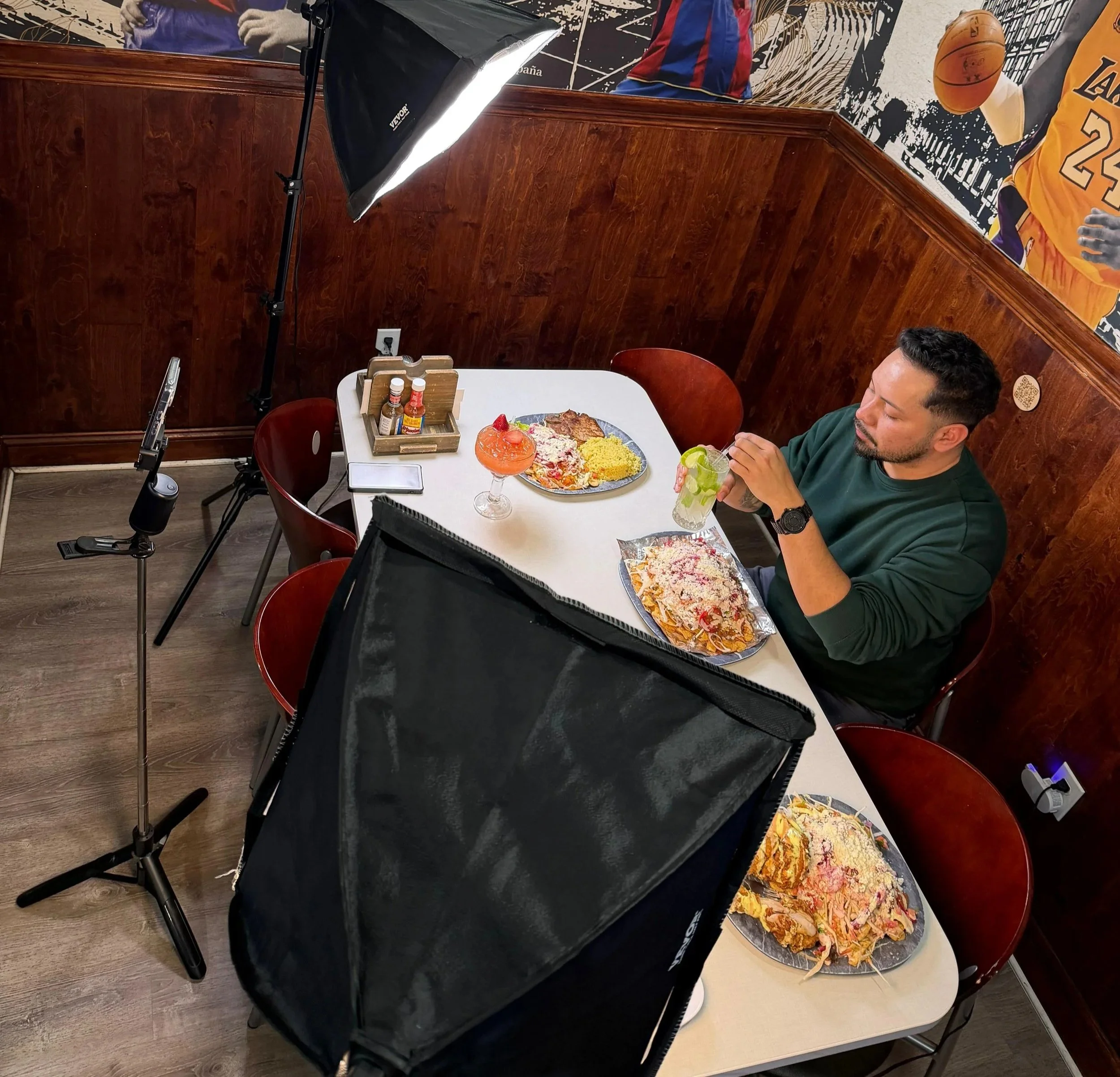 Un hombre comiendo pizza en un restaurante con decoraciones deportivas, como un póster de baloncesto, y varias salsas en la mesa.