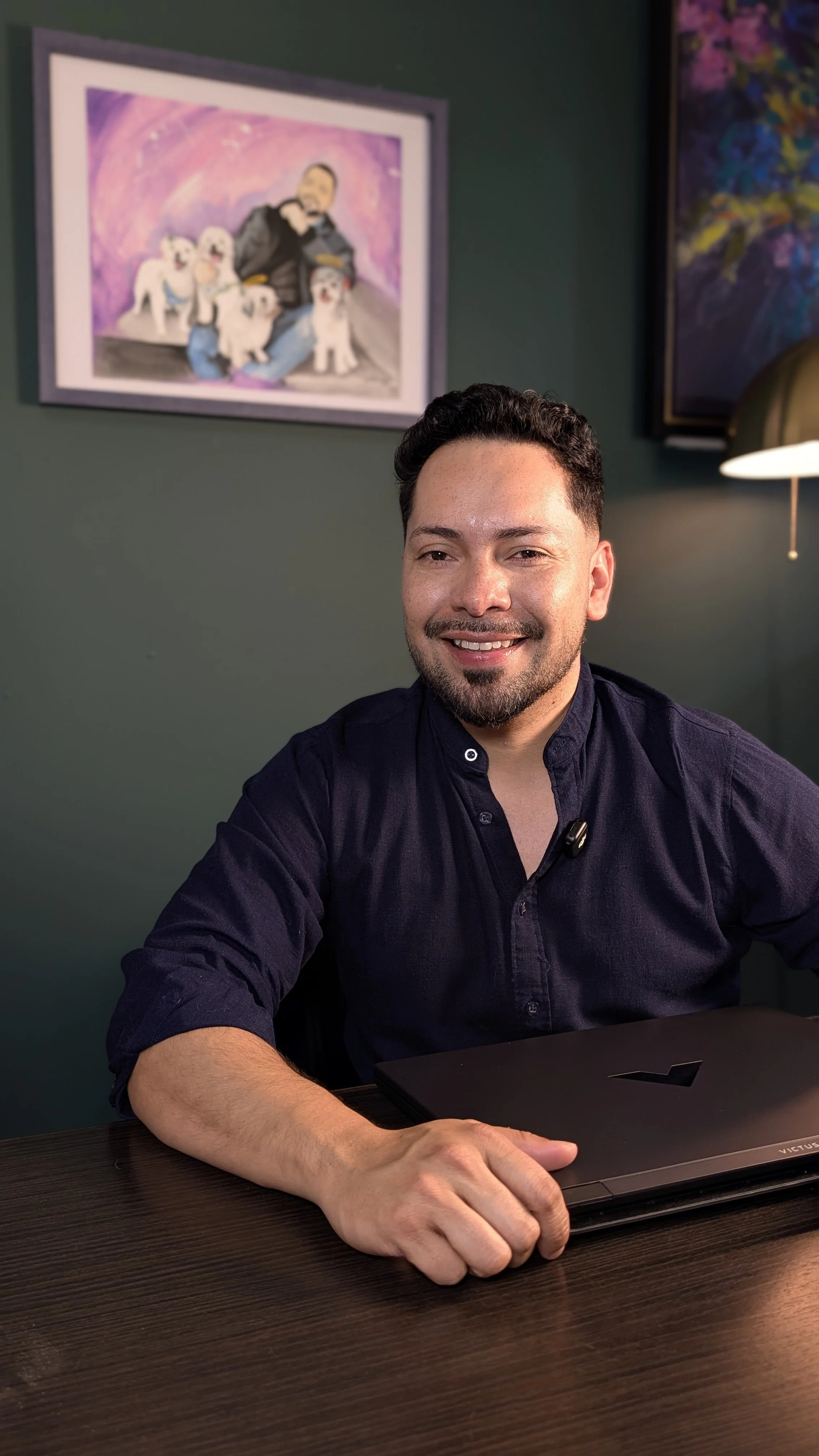Hombre sonriendo sentado en una mesa con una computadora portátil en un ambiente de oficina o estudio, con pinturas abstractas en las paredes.