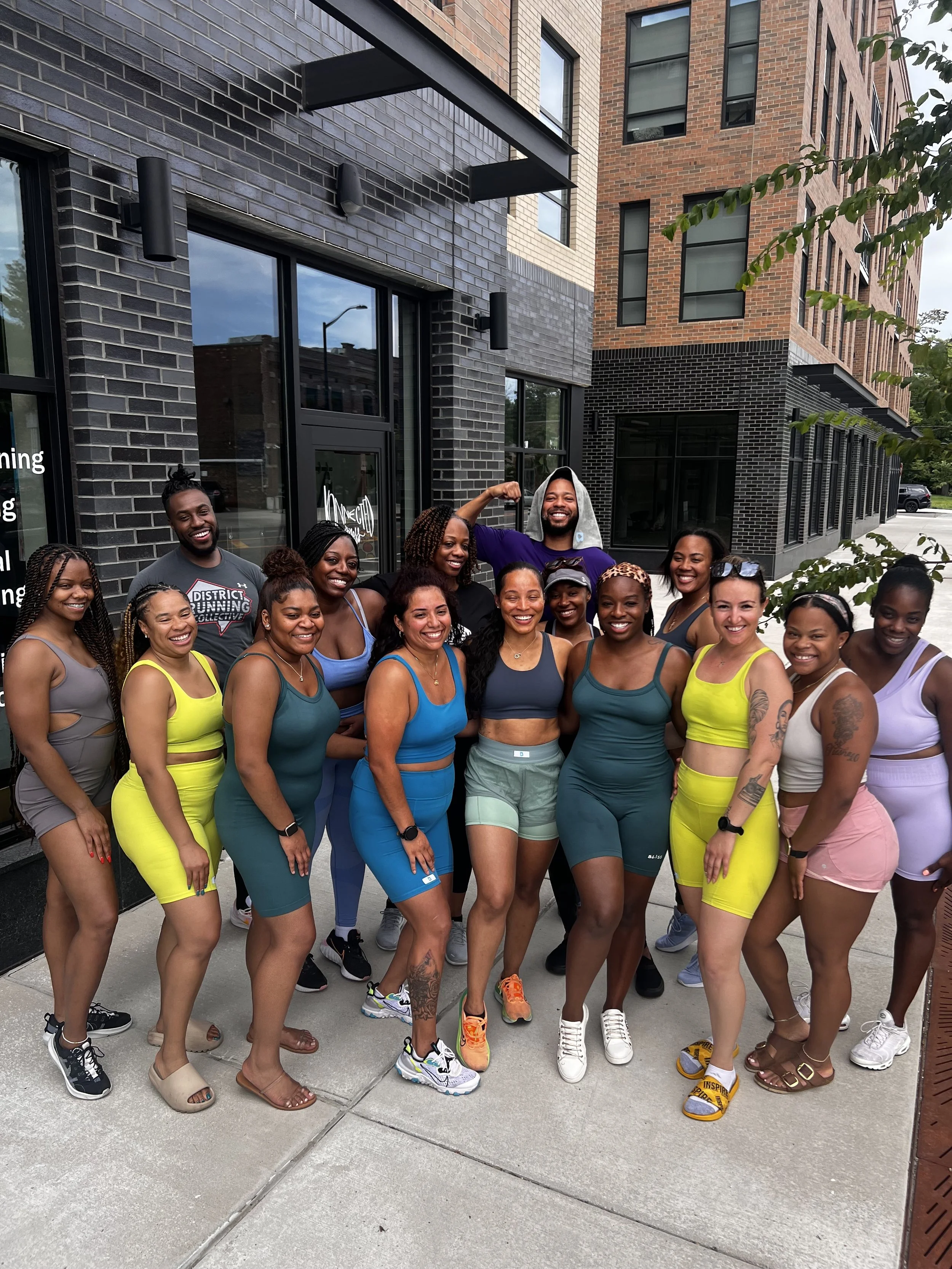 Group of diverse women and men in colorful athletic wear standing outside in front of a modern building, smiling and posing for a photo after a workout.