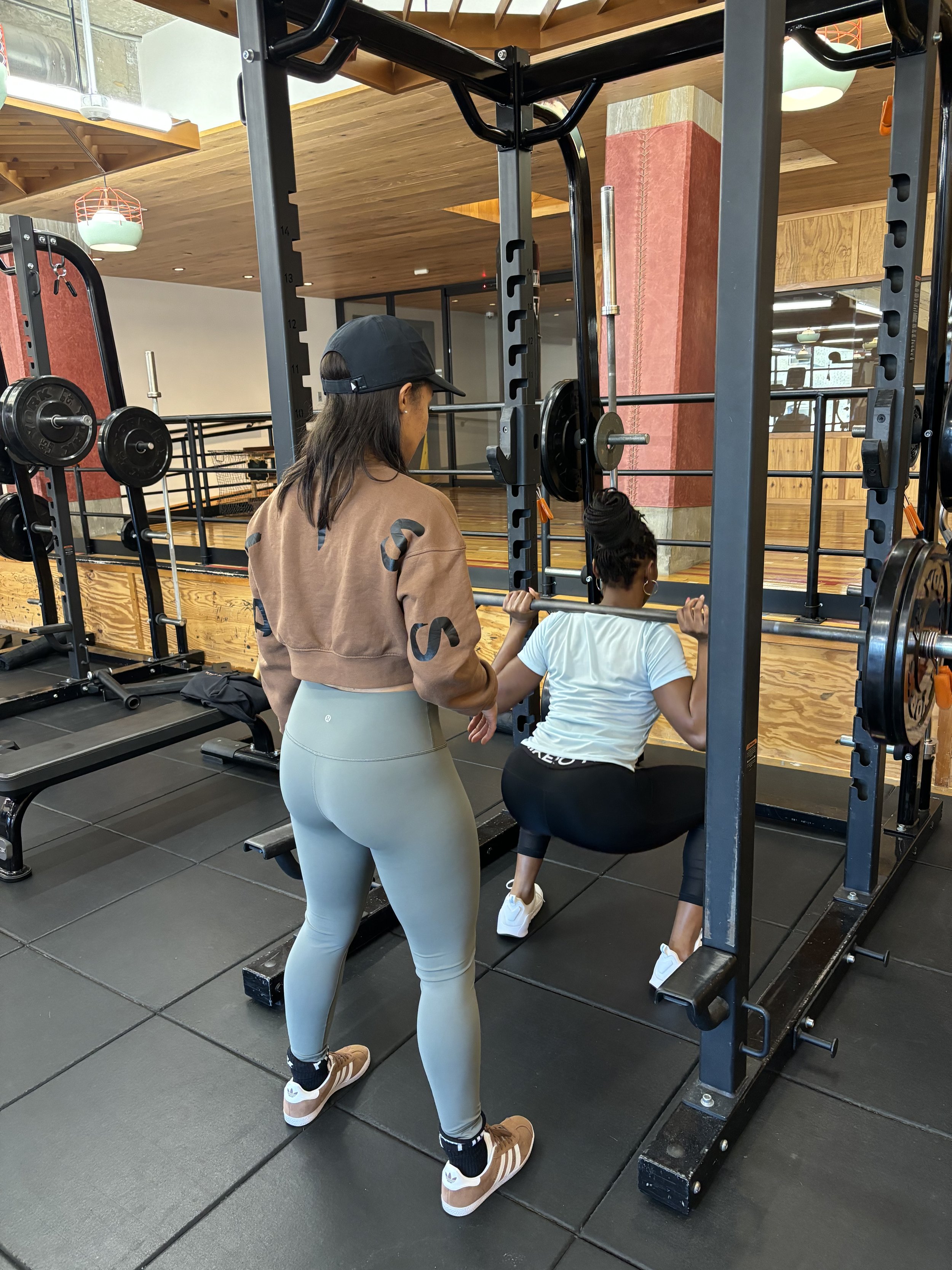 A woman in light gray leggings, a brown sweatshirt, and brown sneakers is standing at a squat rack, helping a woman in a white T-shirt and black leggings with her squats in a gym that has wooden accents and black flooring.