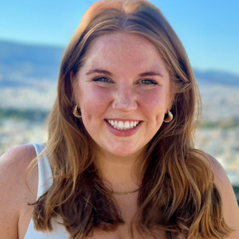 A smiling young woman with long, wavy red hair, outdoors with the Athenian cityscape and mountains in the background.
