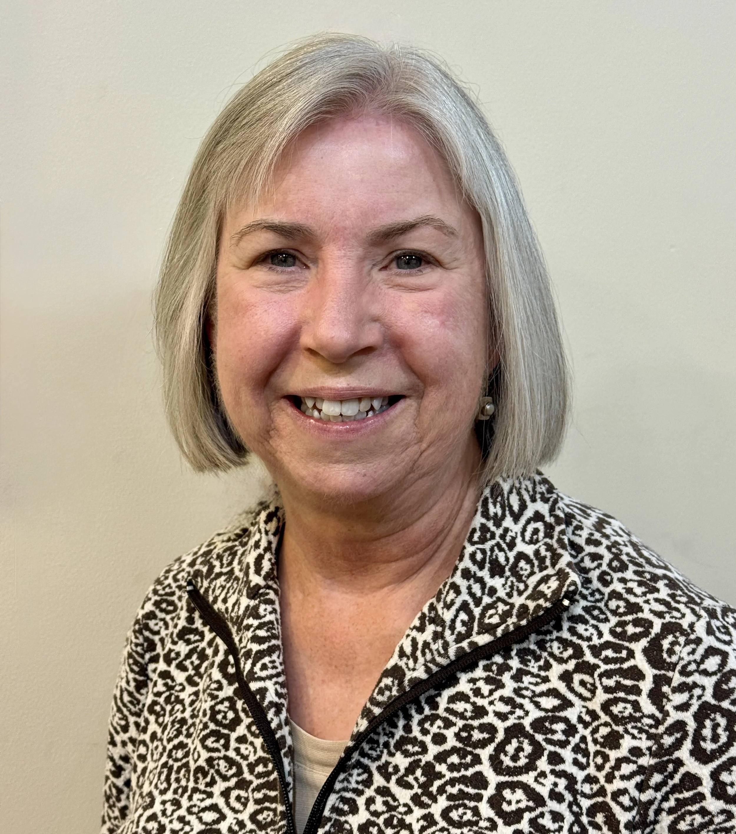 Close-up of smiling middle-aged woman with blonde hair wearing a black and white animal print jacket against a plain light-colored wall.
