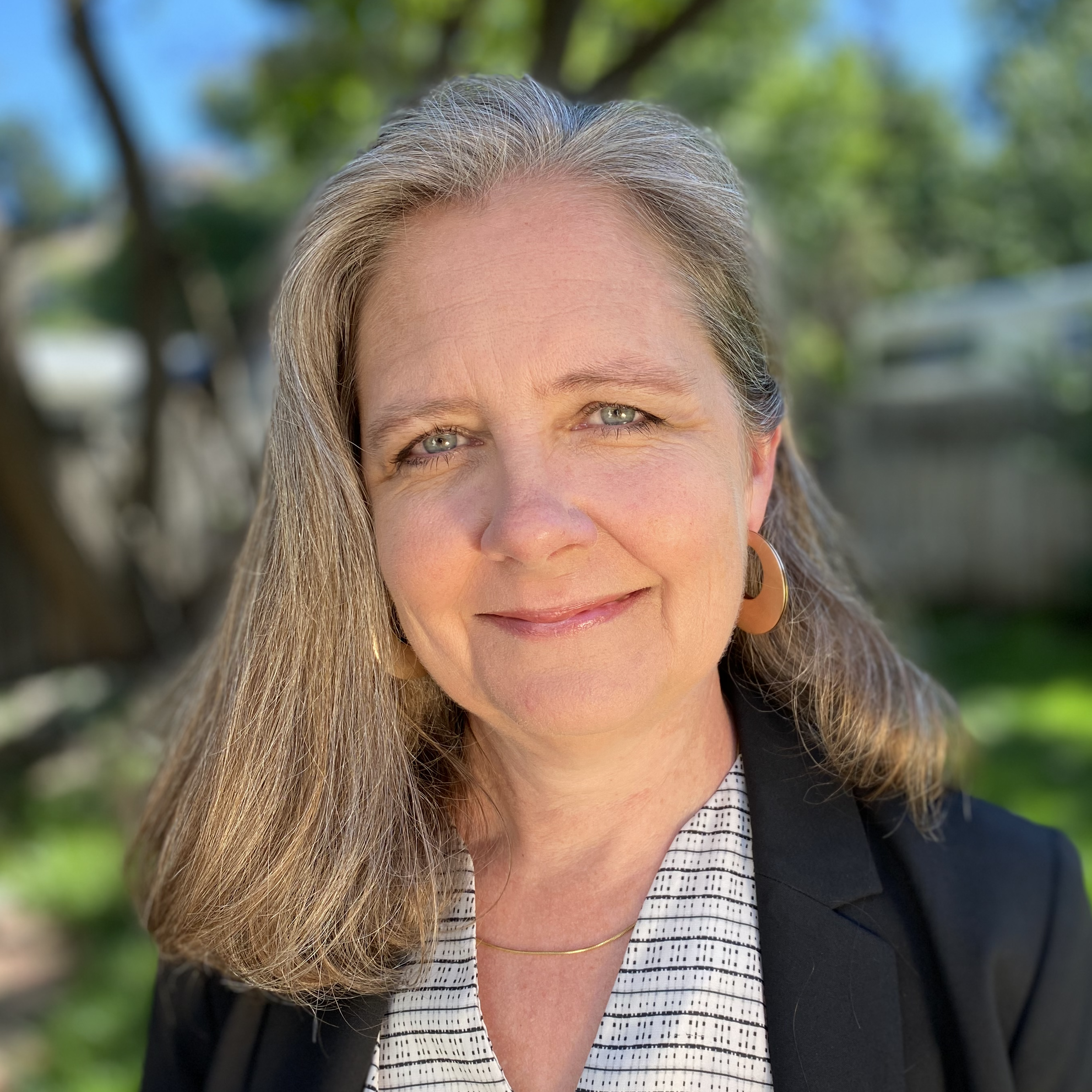 Close-up of a woman with gray hair, blue eyes, smiling outdoors with green trees and blue sky in the background.