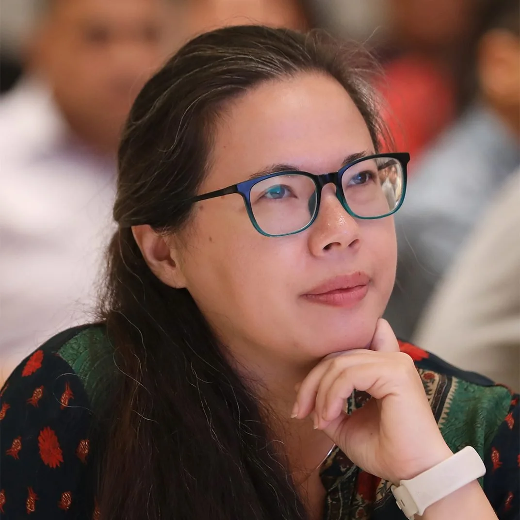 A woman with glasses resting her chin on her hand, attending an event with a blurred crowd in the background.