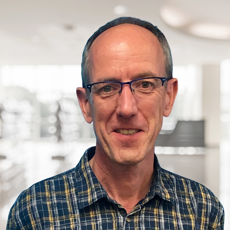 Portrait of a middle-aged man with short dark hair, wearing glasses and a blue plaid shirt, smiling in an indoor setting with a blurred background.