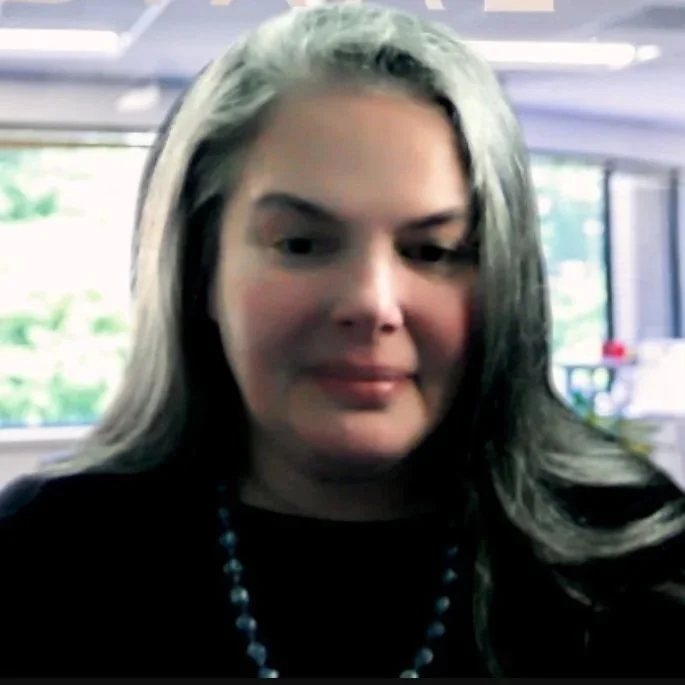A woman with long gray hair smiling in a bright indoor setting with large windows in the background.