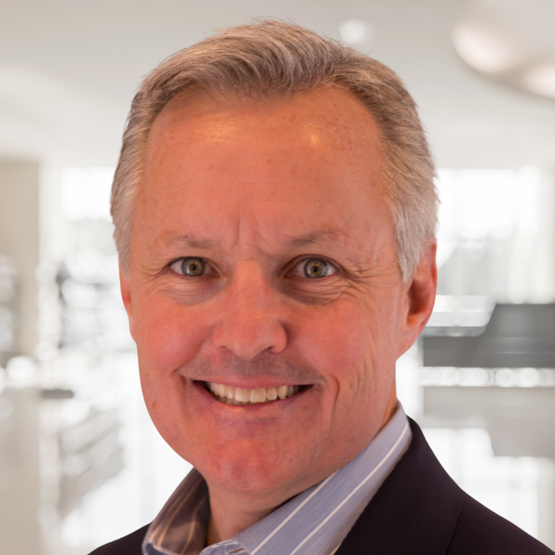 A middle-aged man with short, gray hair, smiling, dressed in a dark blazer and light shirt, in a bright, modern indoor setting.