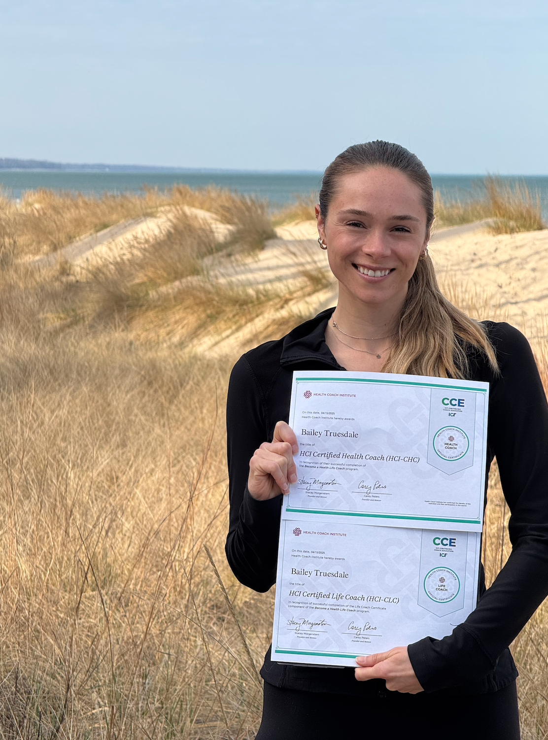 A woman smiling at the camera, holding two certification diplomas, standing on a sandy coastal area with tall grass and the ocean in the background.