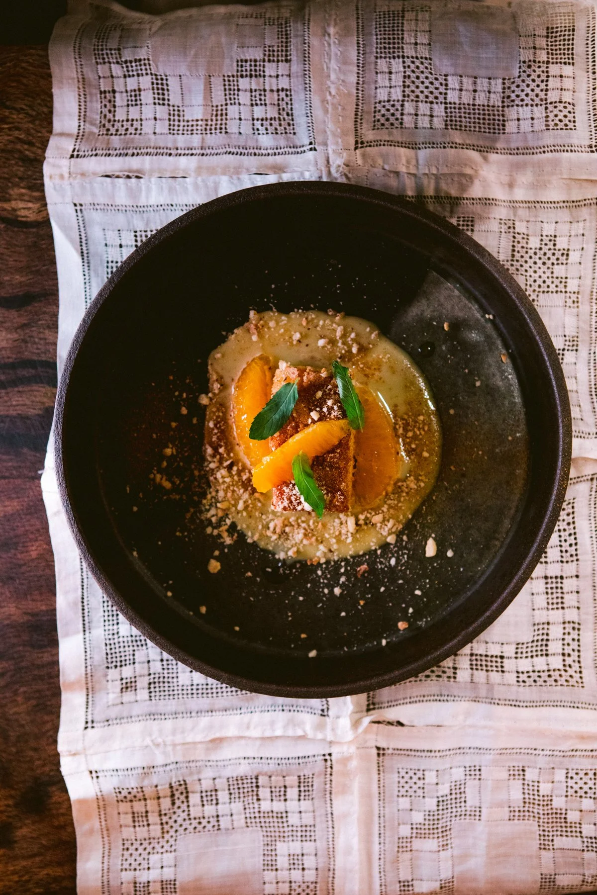 Citrus olive oil cake with burnt citrus cream, orange slices, lemon verbena syrup, hazelnut, and persimmon powder sits in a black ceramic bowl on a lace cloth draped over dark wood.