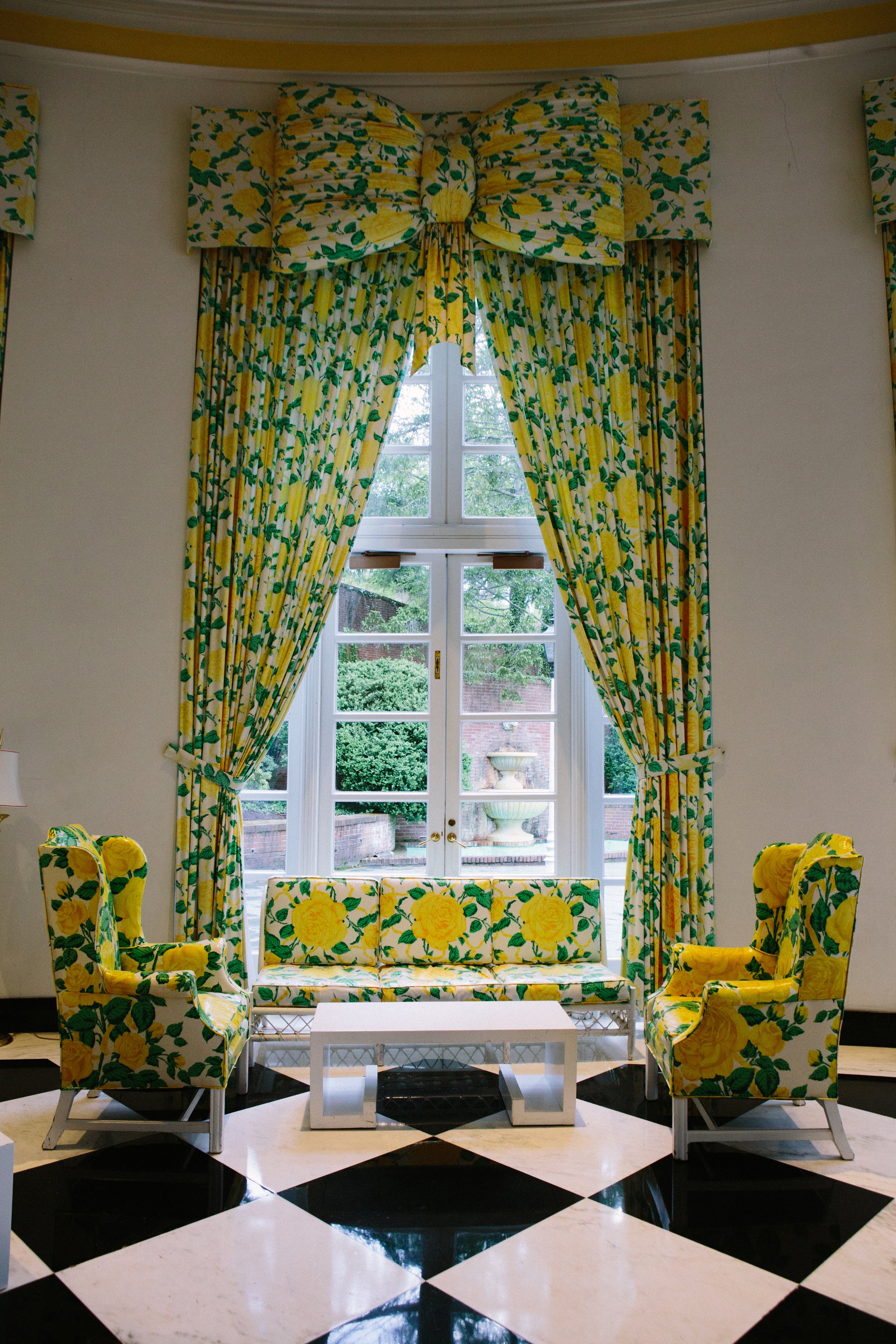 A living room window with a large yellow floral-patterned curtain and matching chairs, white table, and a black-and-white checkered tile floor.