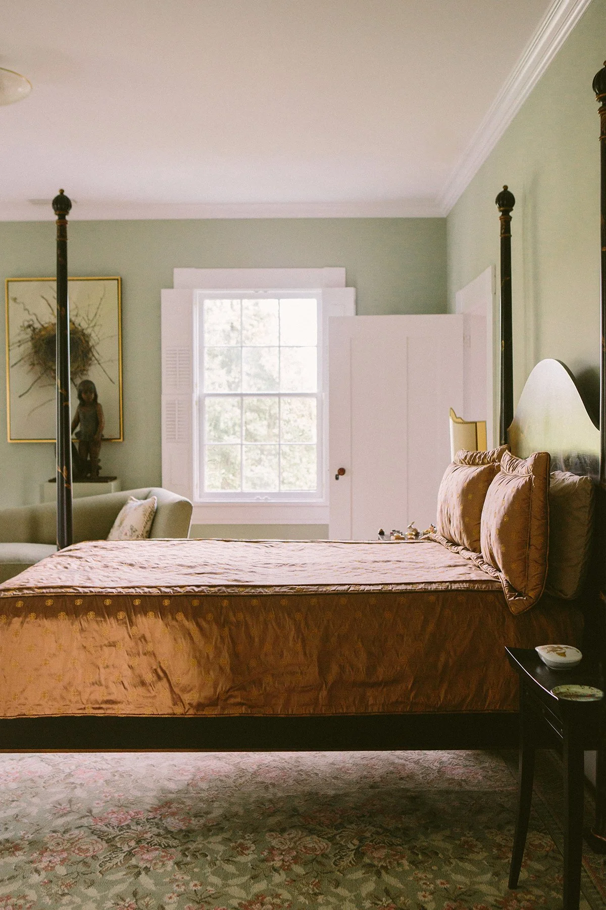 A bedroom with an antique four-poster bed covered in a gold-colored patterned bedspread & matching pillows, a window w/ white shutters, a green wall, a dresser with decorative items, and a vintage floral area rug at a private estate in Lancaster Ohio