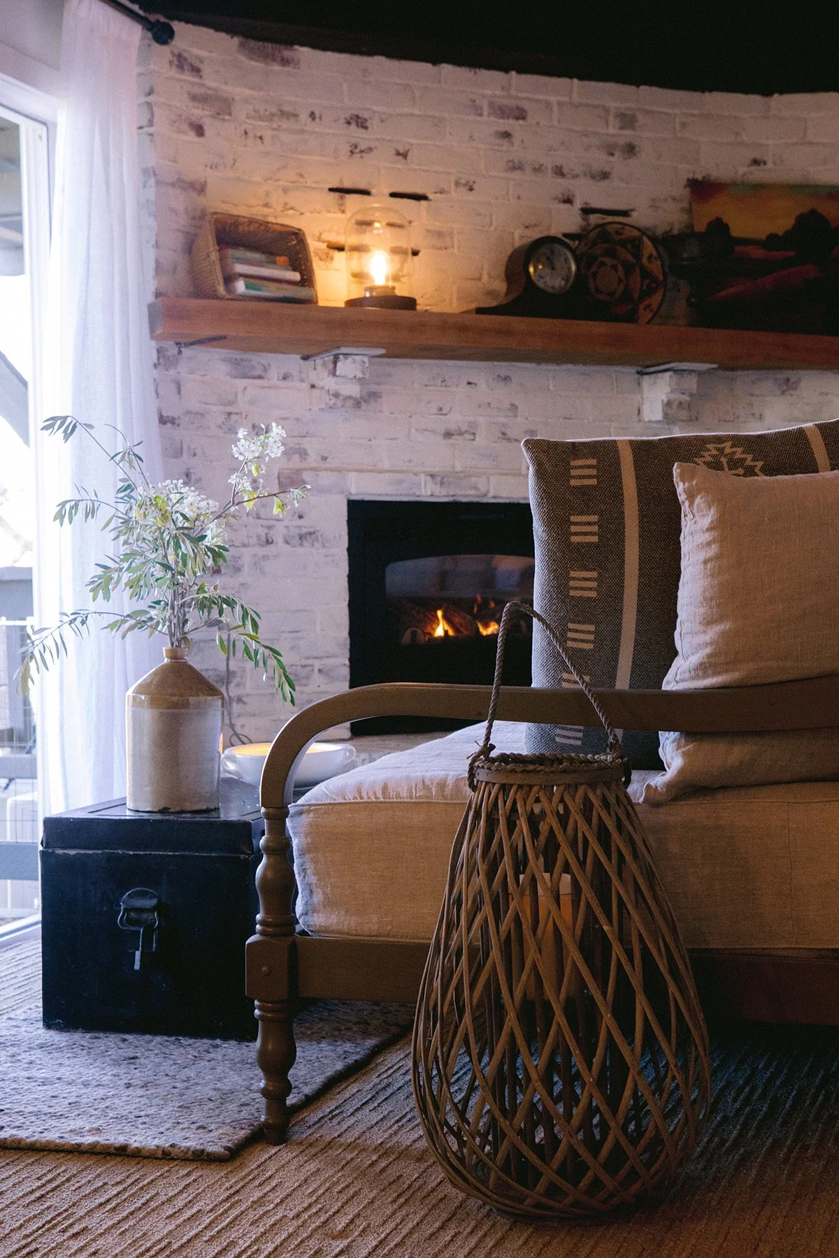 Cozy living room with a white brick fireplace, lit, and a wooden mantel top decorated with books, a framed picture, and a vintage lantern. A beige armchair with patterned pillows, a dark side table with a white vase and green plant, and an empty wicker basket in the foreground.