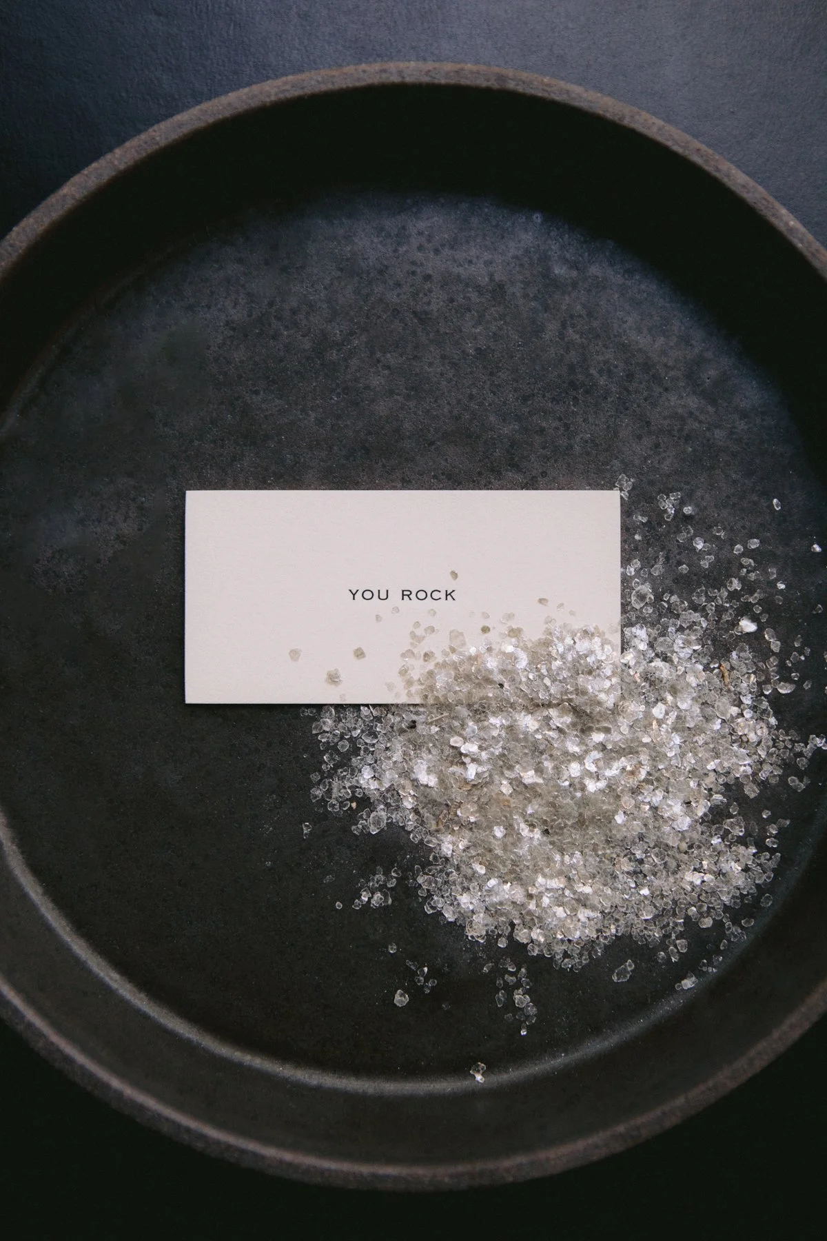 A black round tray with a white card that says "YOU ROCK" and scattered small, clear, and reflective crystals.