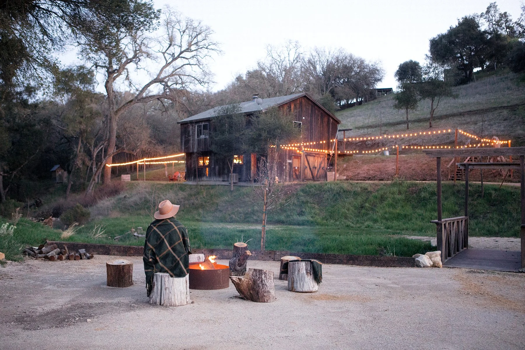 A person wearing a large hat and plaid blanket, sitting by a campfire surrounded by tree stumps, at dusk in a rural outdoor setting with a rustic wooden barn and string lights on a hillside in the background.