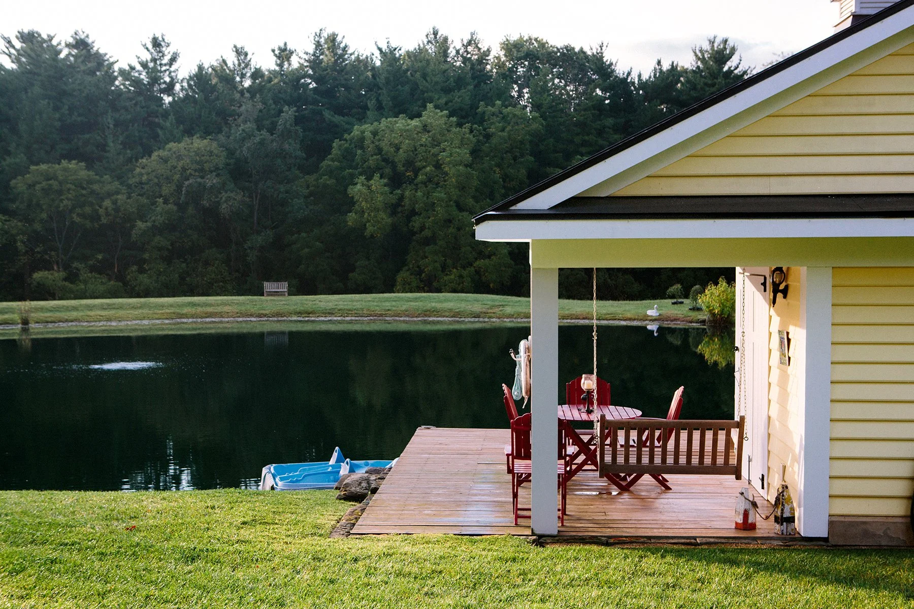 Yellow lake house on a private estate with a porch overlooking a pond with a wooden deck, red chairs, and a small paddle boat on the water, surrounded by green trees, in Lancaster Ohio.