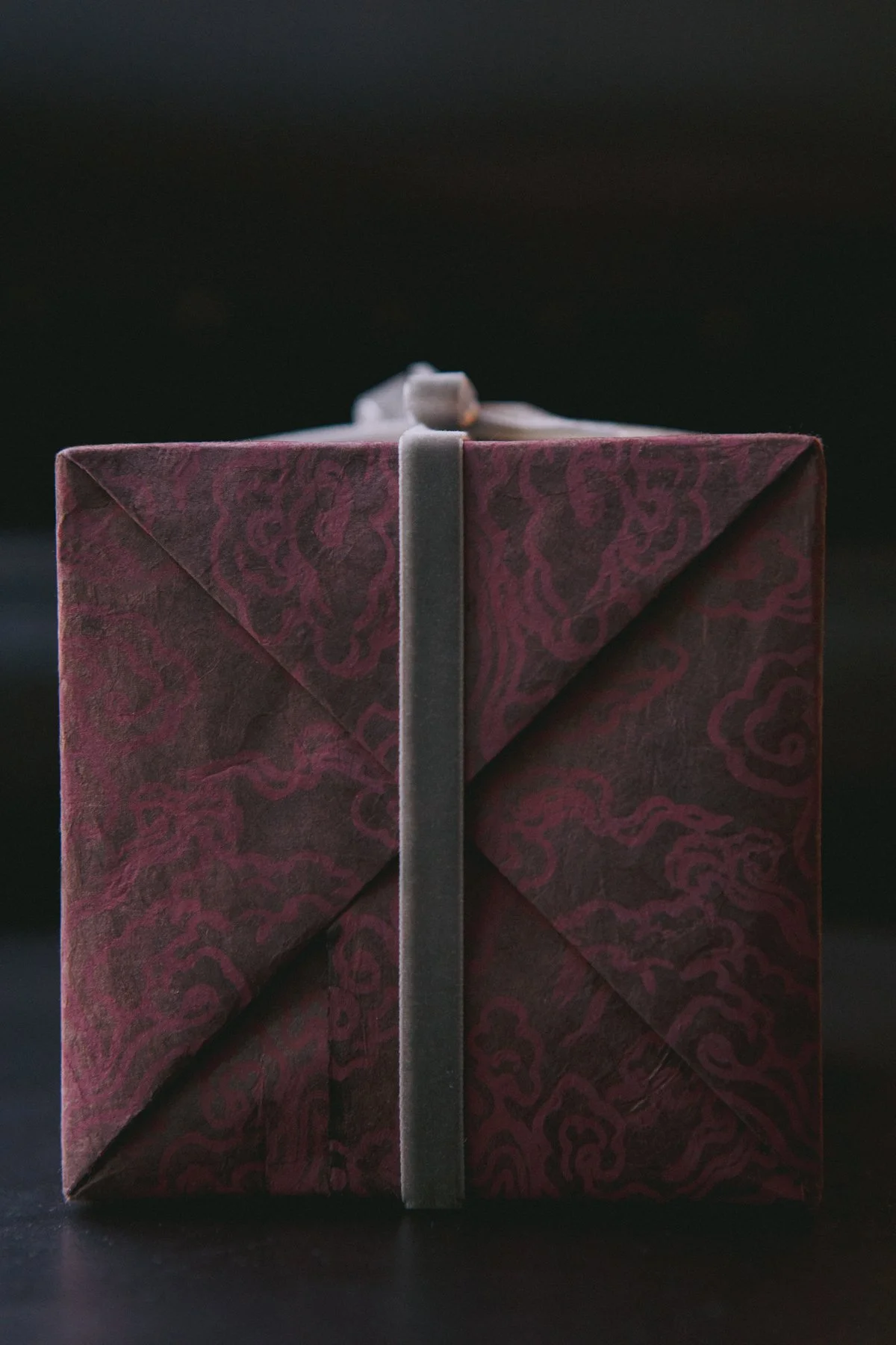 A close-up photograph of a gift box wrapped in dark red paper with a subtle ornate pattern, tied with a gray velvet ribbon, and topped with a small bow, against a black background.