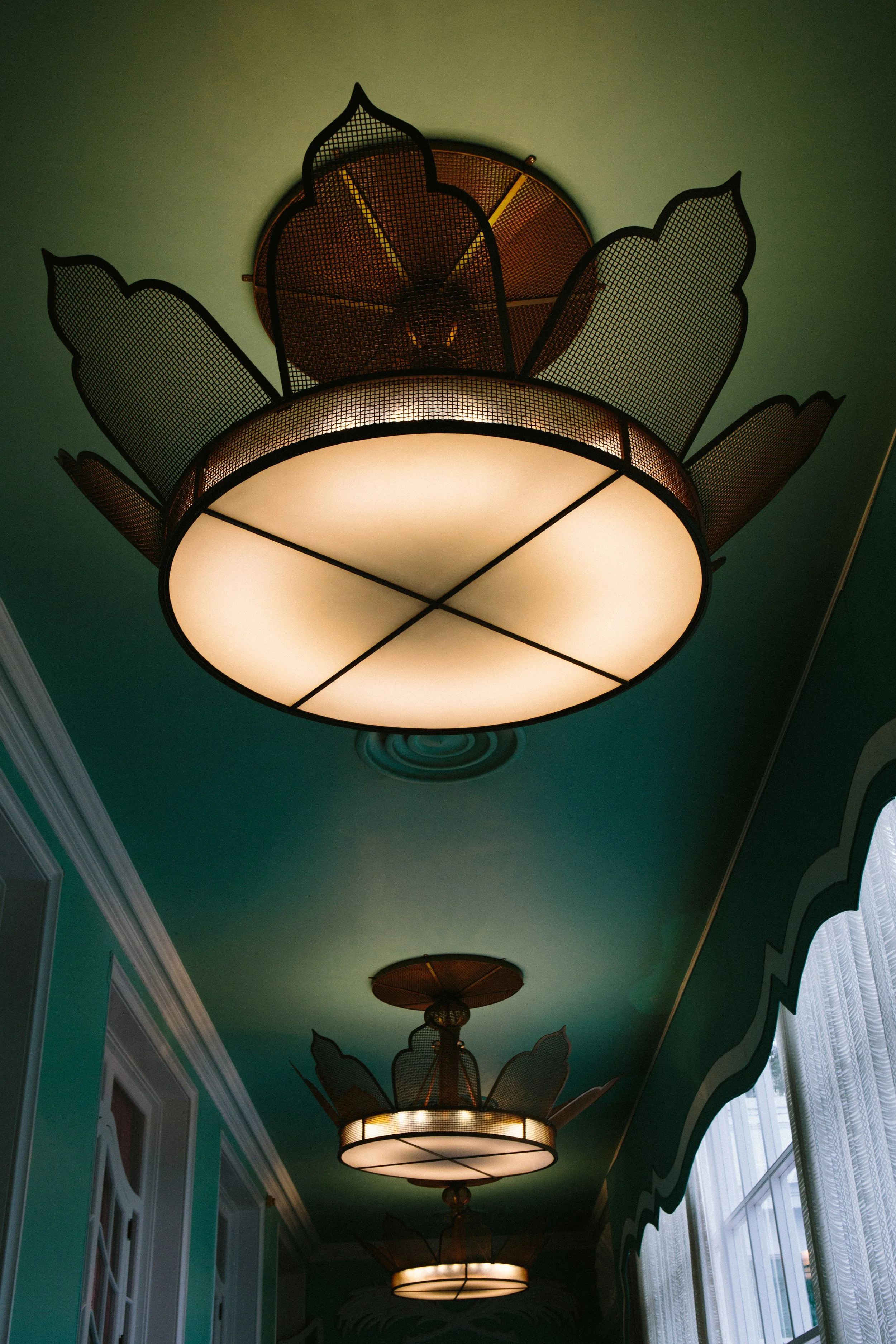 A ceiling with multiple ornate hanging light fixtures, with a green painted ceiling and windows with sheer white curtains on the side.