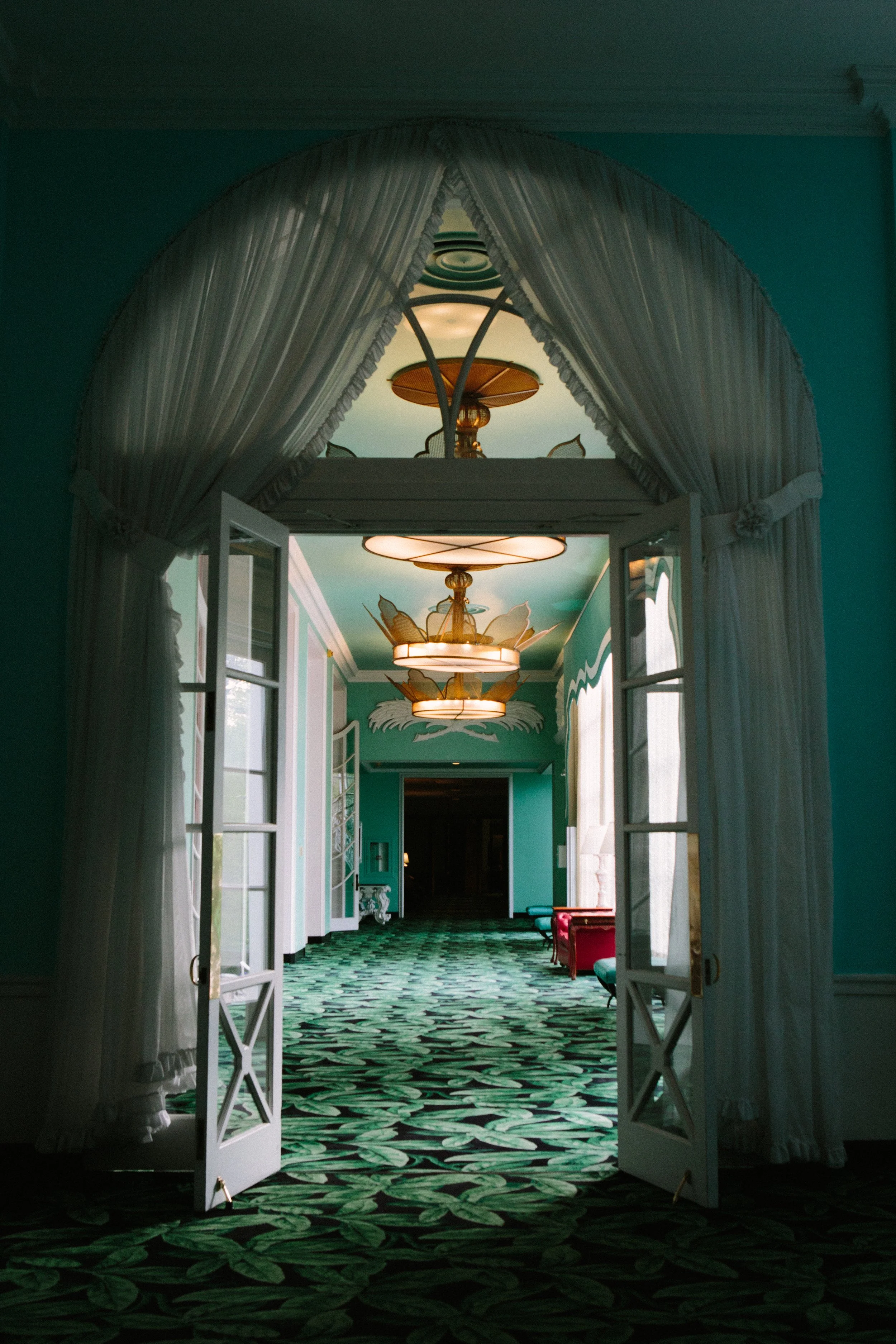View of a decorated hallway seen through open white French doors with curtains, featuring green walls, patterned green carpet, and ornate ceiling light fixtures.