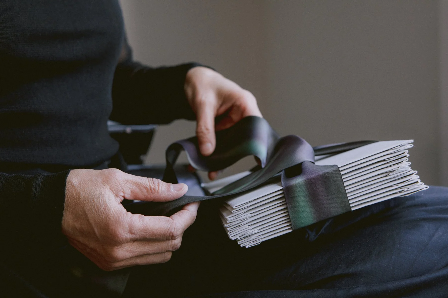 Close-up of a person's hands holding a stack of papers, tied with a black ribbon, with a dark-colored ribbon also being placed on top.