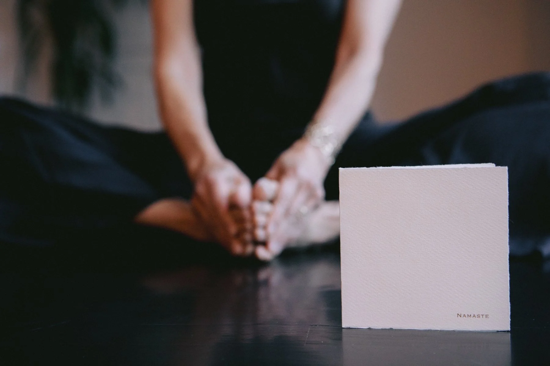 Person sitting cross-legged with hands in namaste position on a dark surface, in front of a white card with 'Namaste' printed on it.
