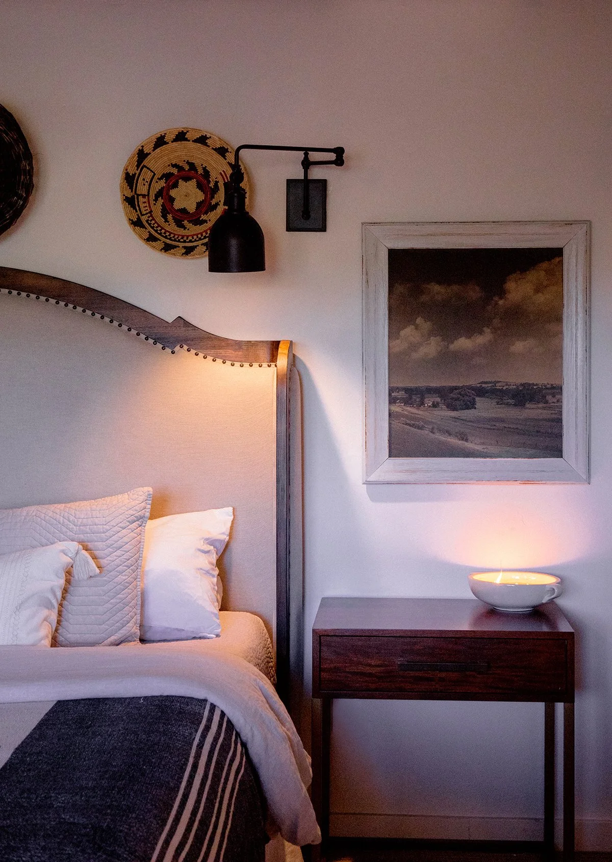A bedroom corner featuring a bed with white pillows and a beige headboard, a wooden nightstand with a lit ceramic bowl, a framed landscape painting, and wall decor including a woven circular art piece, a black wall-mounted lamp, and a black wall fixture.