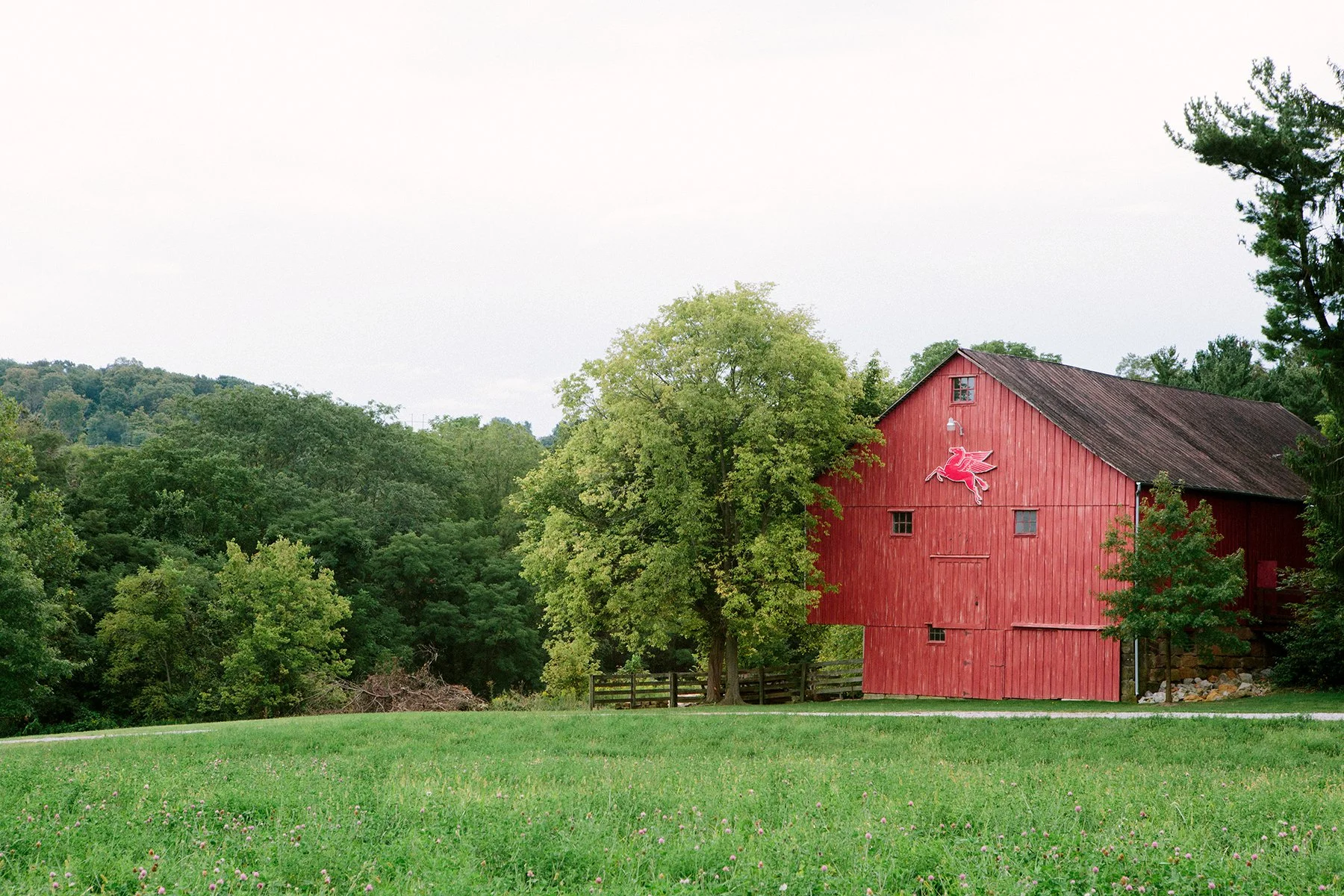 Landscape image of a red barn with a Pegasus emblem, surrounded by green trees and a grassy field under an overcast sky - at a private estate in Lancaster Ohio.