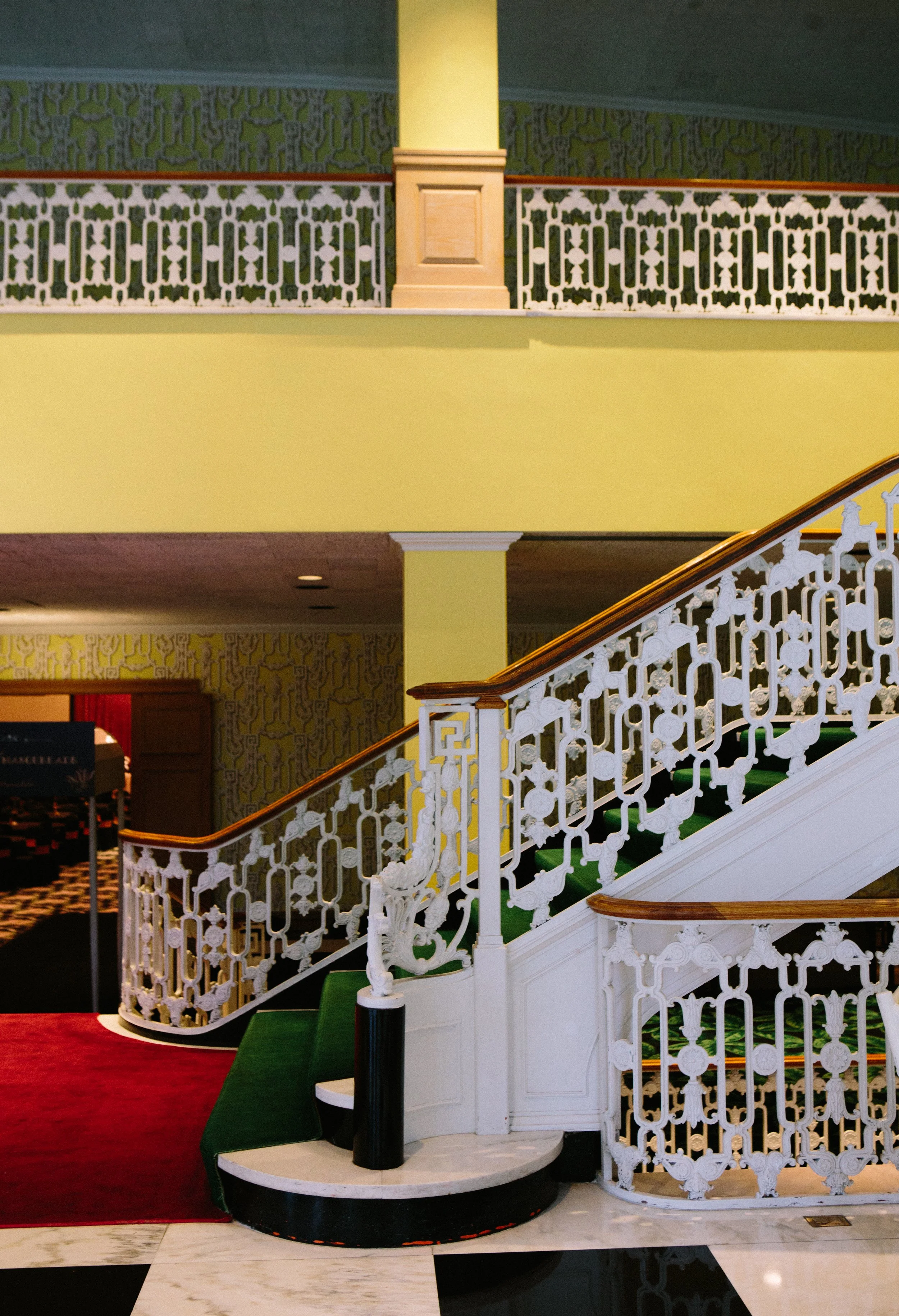 Interior of a building with a decorative white ornate staircase with wooden handrails, green carpeted steps, yellow walls, patterned wallpaper, and a patterned black and white floor.
