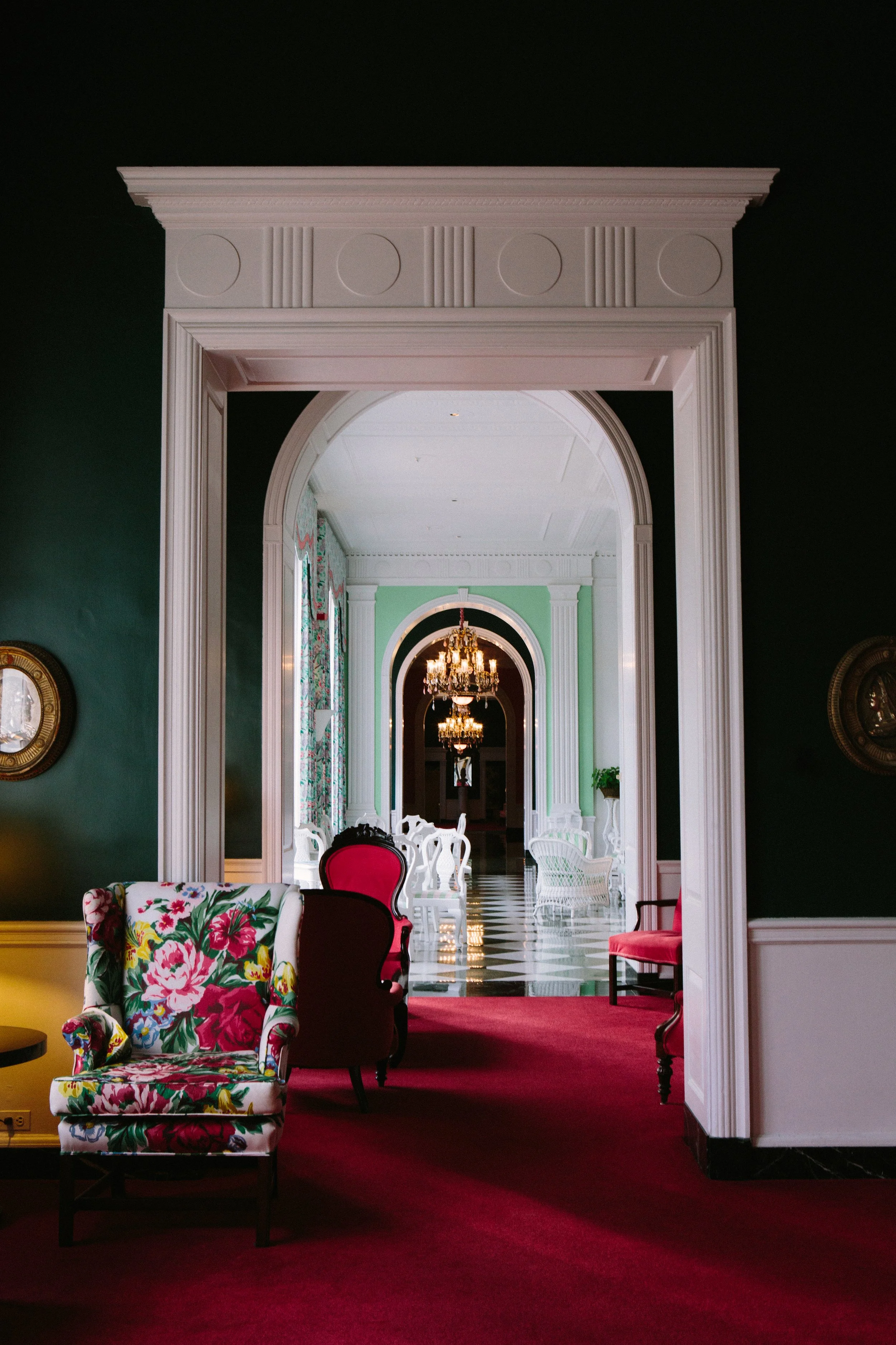 Interior view of a luxurious room with a large doorway leading to a chandelier-lit hallway. The room features vibrant, colorful furniture including a floral armchair, red cushioned chairs, and white chairs. The walls are dark green with a clock on the left and detailed moldings around the doorway. The hallway has a green wall with windows and more chandeliers visible.