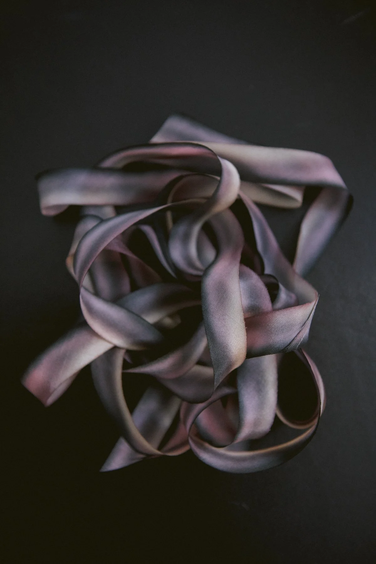 A crumpled, twisted, ribbon-like object with shades of gray, purple, and pink on a dark background.