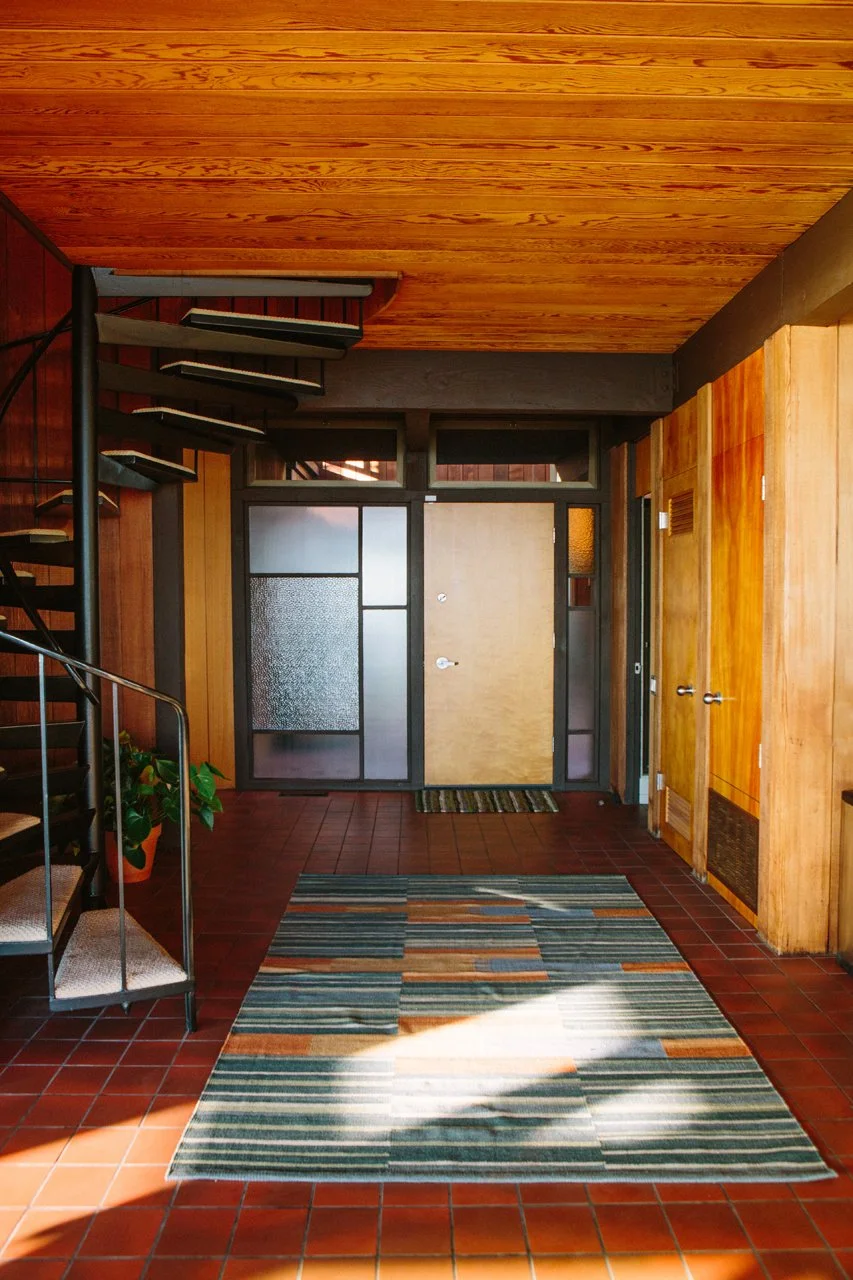 Interior of a 1970's modernist home in Malibu - entryway with a tiled floor, striped rug, wood paneled walls, a glass-paneled front door, and a staircase with black metal railing.