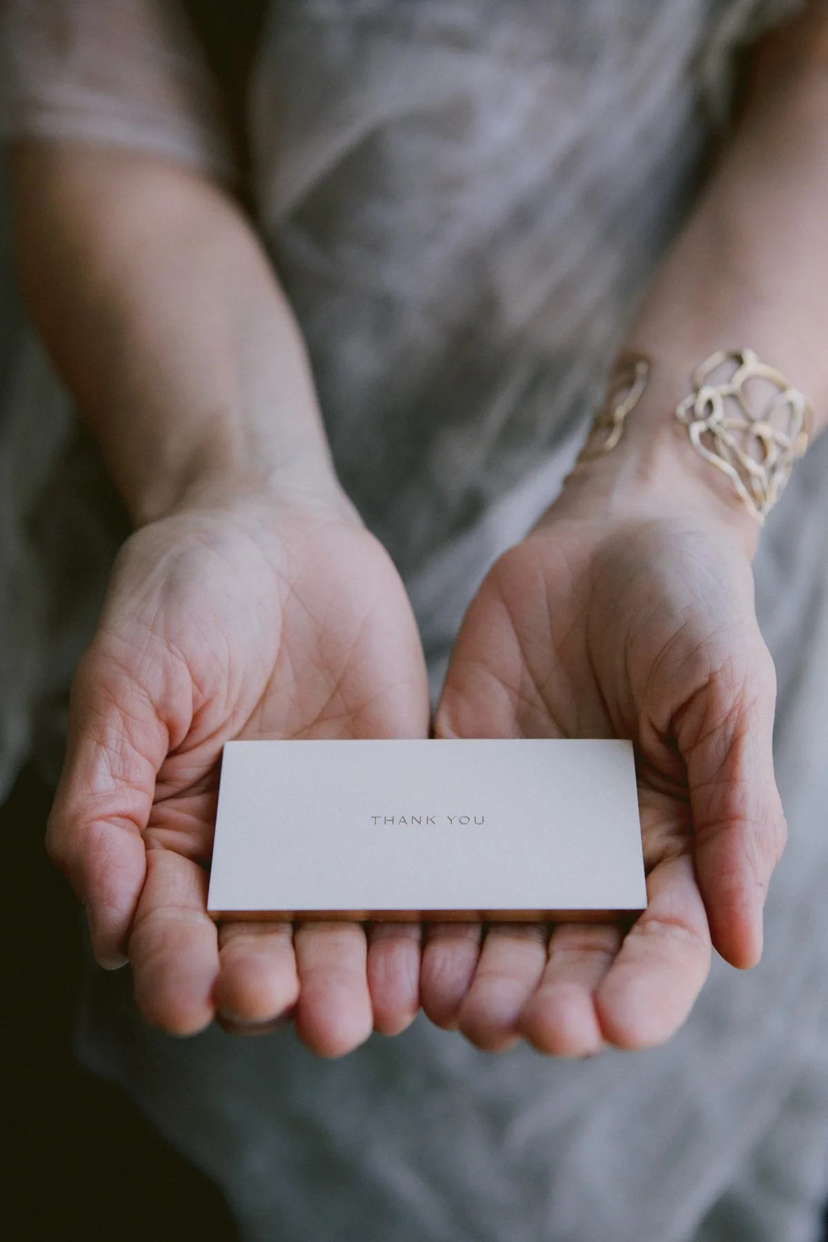 Close-up of elderly person's hands holding a white card with 'Thank You' printed on it, with a young person's hands surrounding the elderly's hands, and a blurred gray shirt in the background.