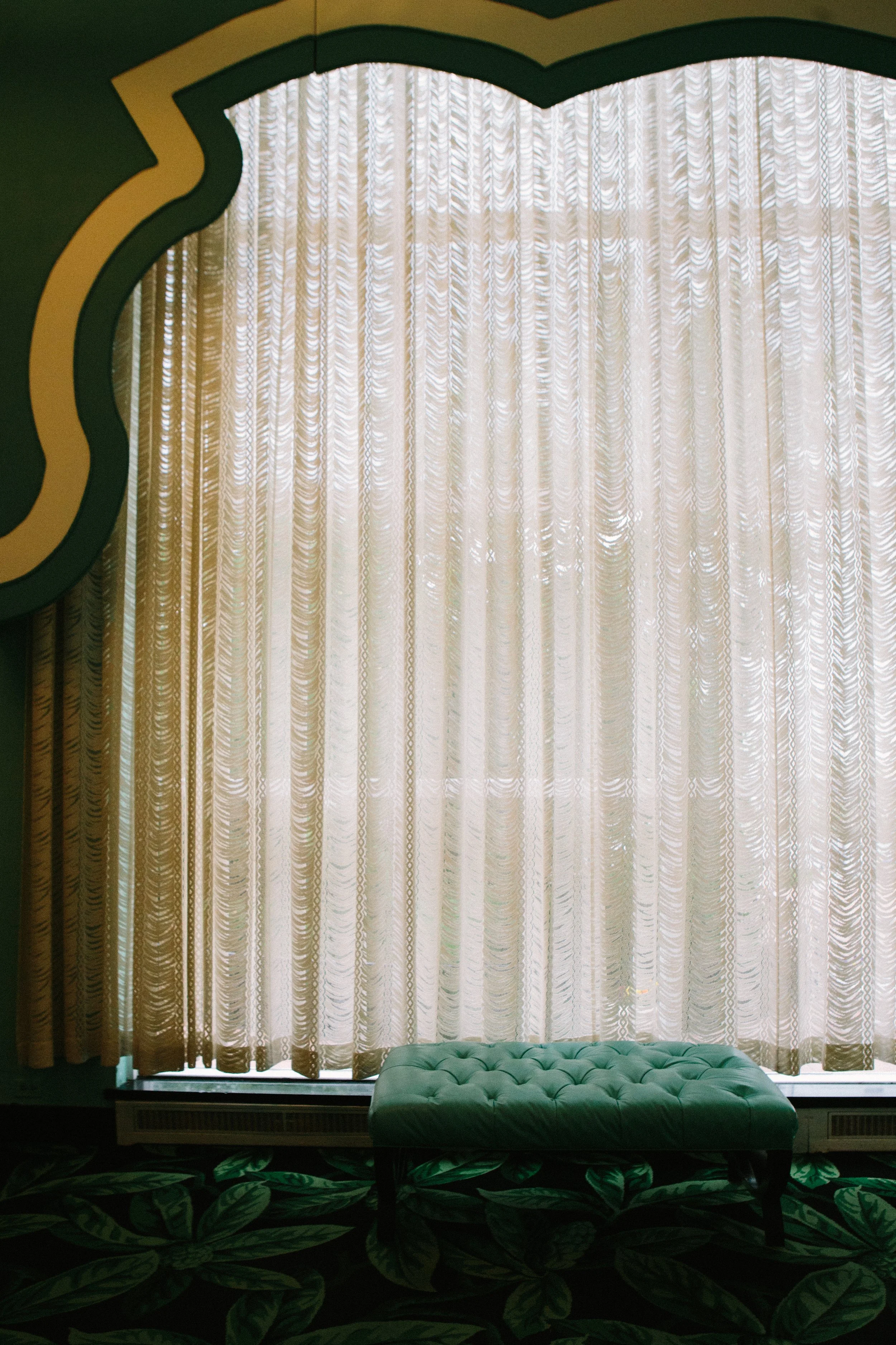 A large window with white patterned curtains and a tufted green bench in front of it. The window is framed by a curved, green and yellow arch.