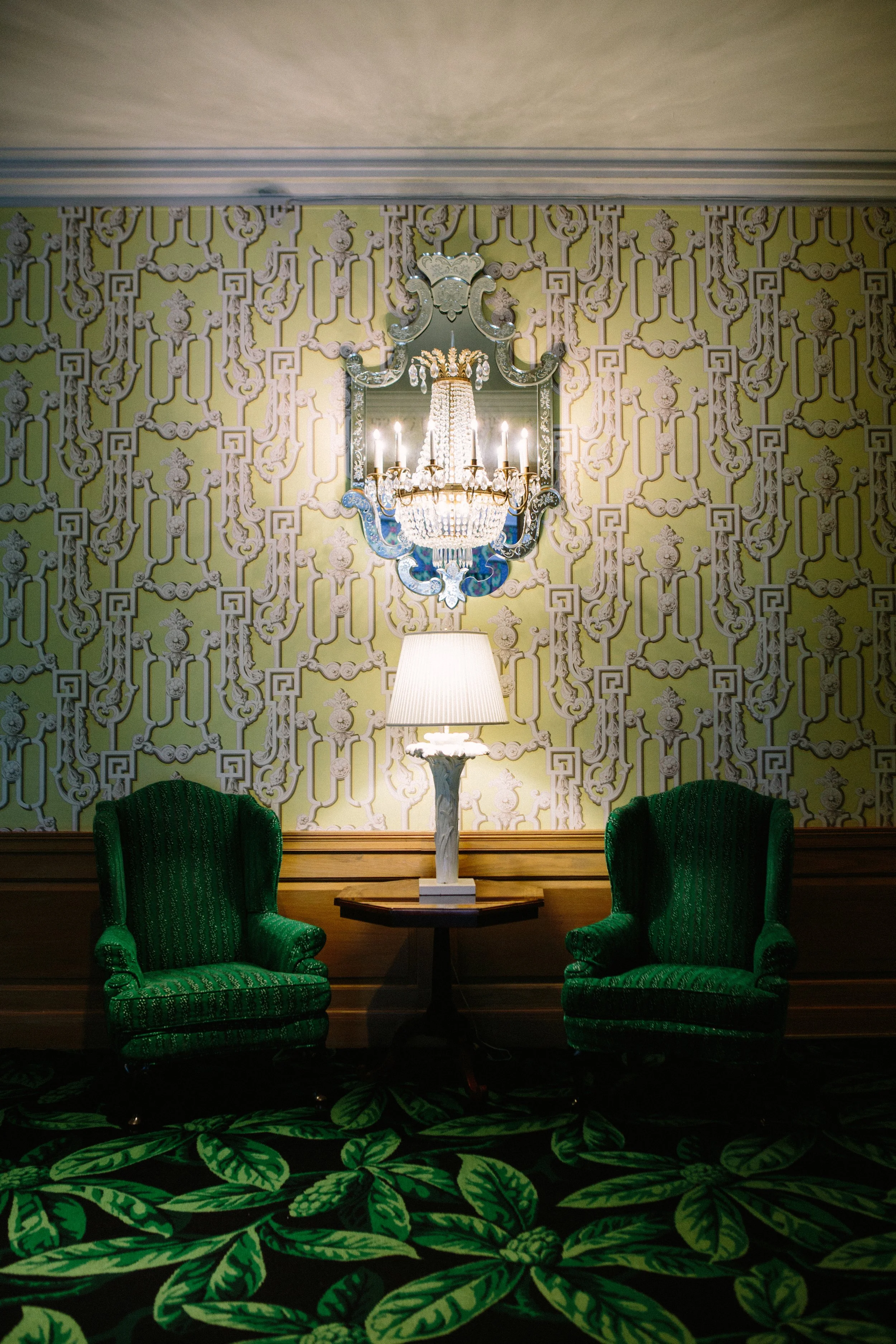 A room with green velvet armchairs, a tall white table lamp, an ornate mirror, a chandelier reflected in the mirror, patterned wallpaper, and a carpet with a green leafy design.