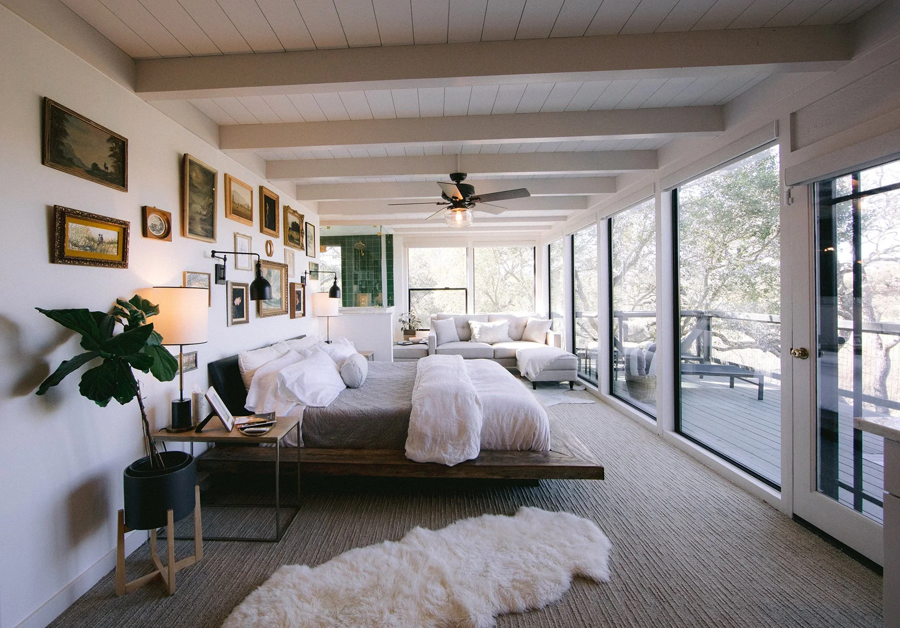Bright living room with white walls, large glass windows, and a sliding door to a balcony. Features a gray bed, white cushions, black and gold framed artwork on the wall, and indoor plants. A ceiling fan with lights, black wall-mounted lamps, and lamps on side tables add lighting. Light-colored carpet and a white fluffy rug complete the decor.