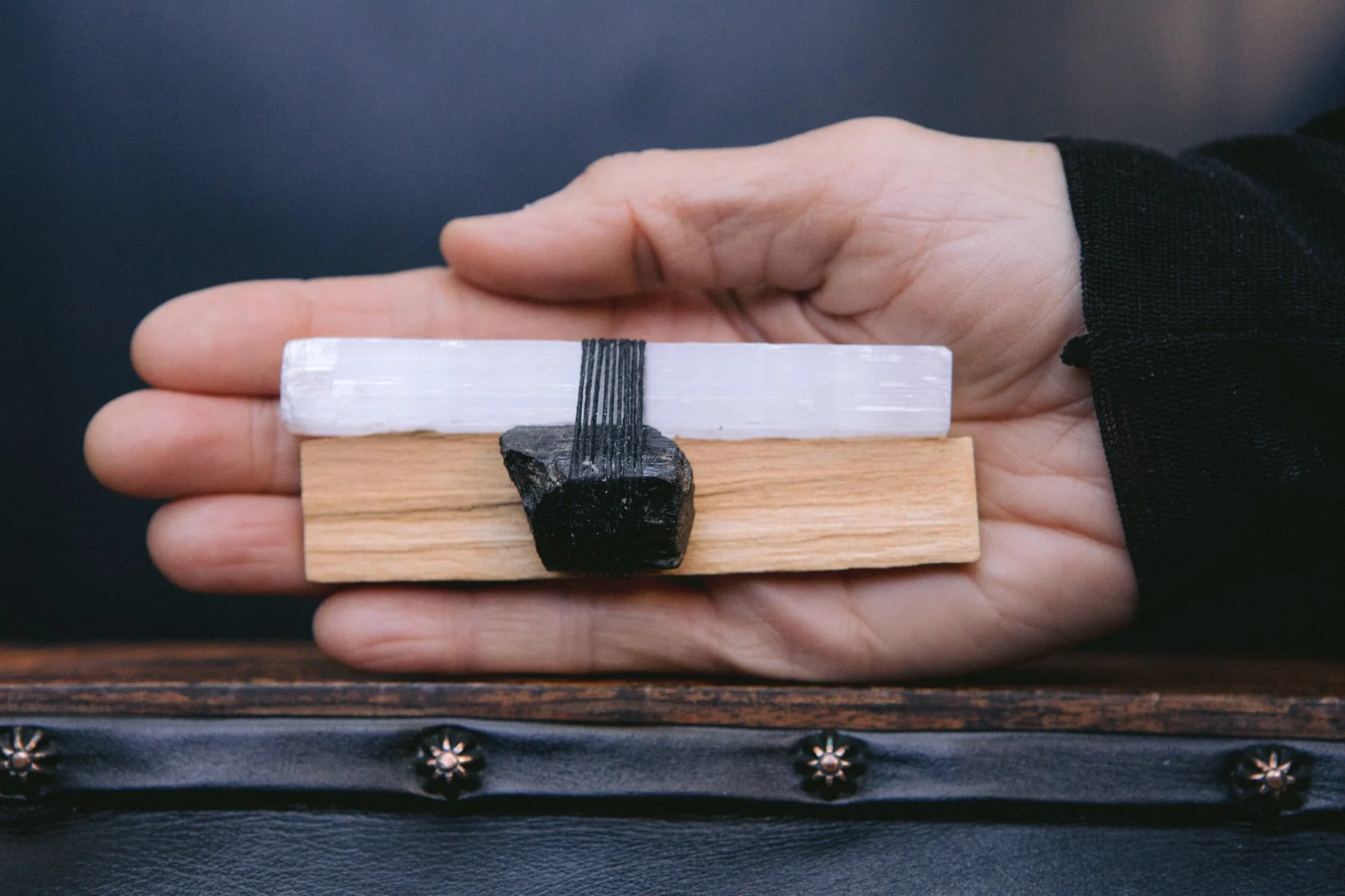 A hand holding three small, rectangular blocks of different wood types stacked in a row, with a black rock and elastic bands on top, on a black surface.