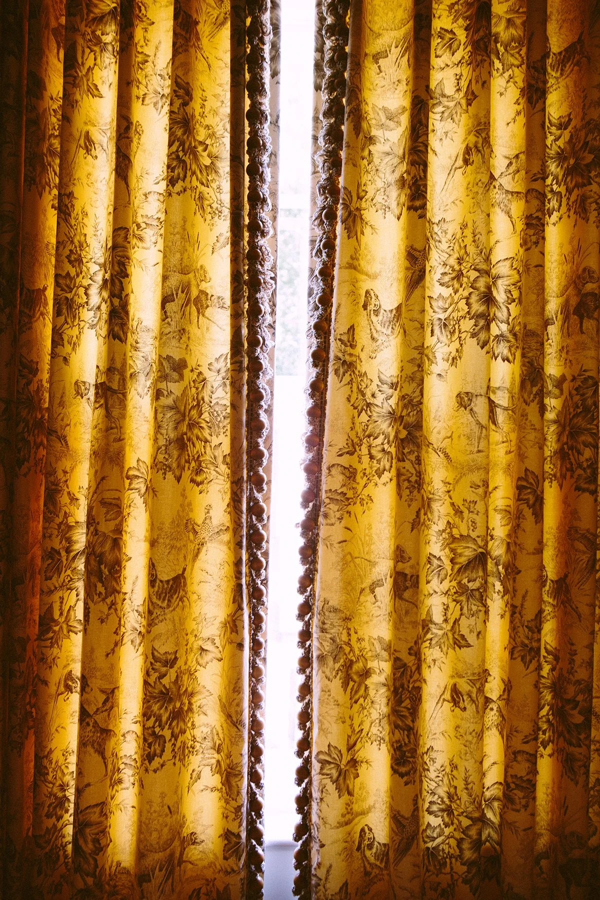 Yellow toile curtains with a decorative tasseled border in front of a window, with light coming through. At a private estate in Lancaster Ohio.