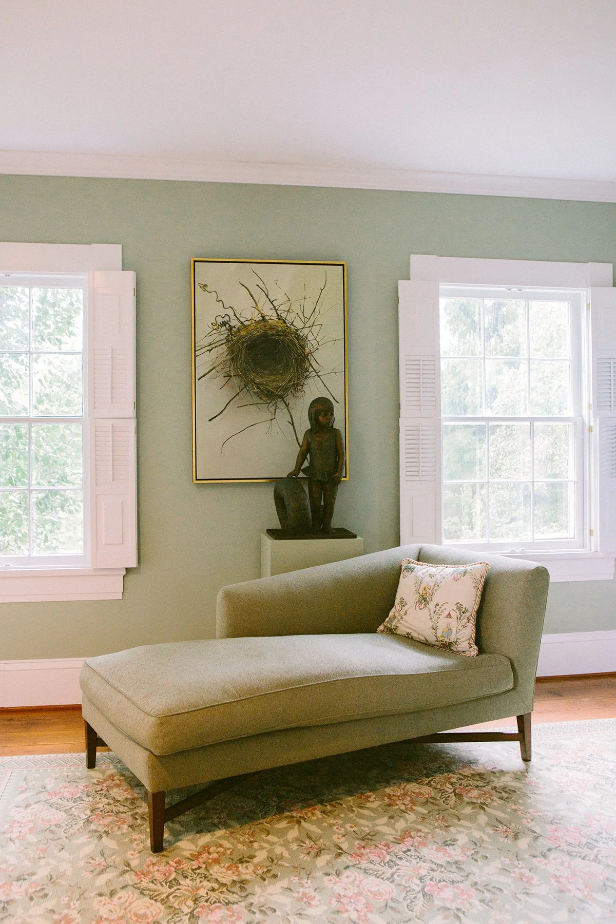 A cozy corner of a private estate in Lancaster Ohio, with a chaise lounge, floral pillow, and a window with white shutters. A large framed photograph of a nest is on the wall, and a black stone sculpture of a girl, on a pedestal.