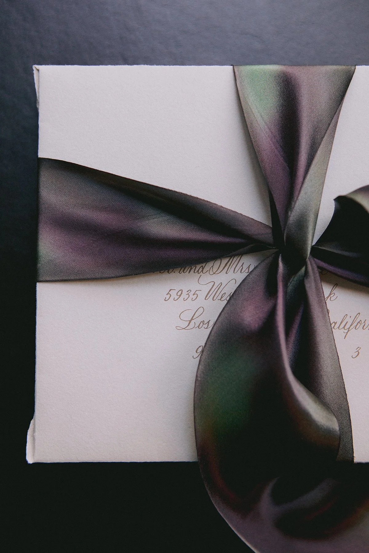 A white wedding invitation envelope with elegant cursive writing, decorated with a dark, multi-colored satin ribbon tied in a bow.