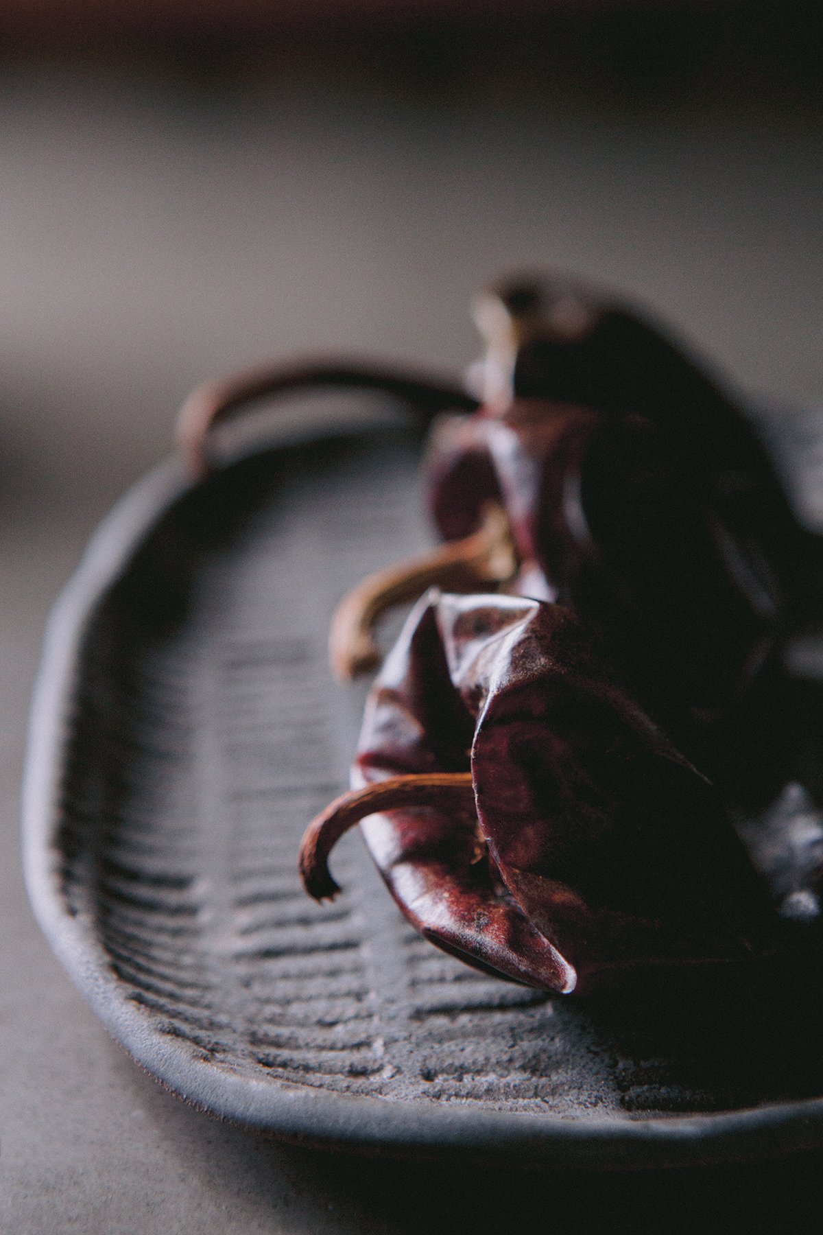 Close-up of three dried red chili peppers , resting on a textured, dark gray ceramic plate against a medium-grey blurred background.