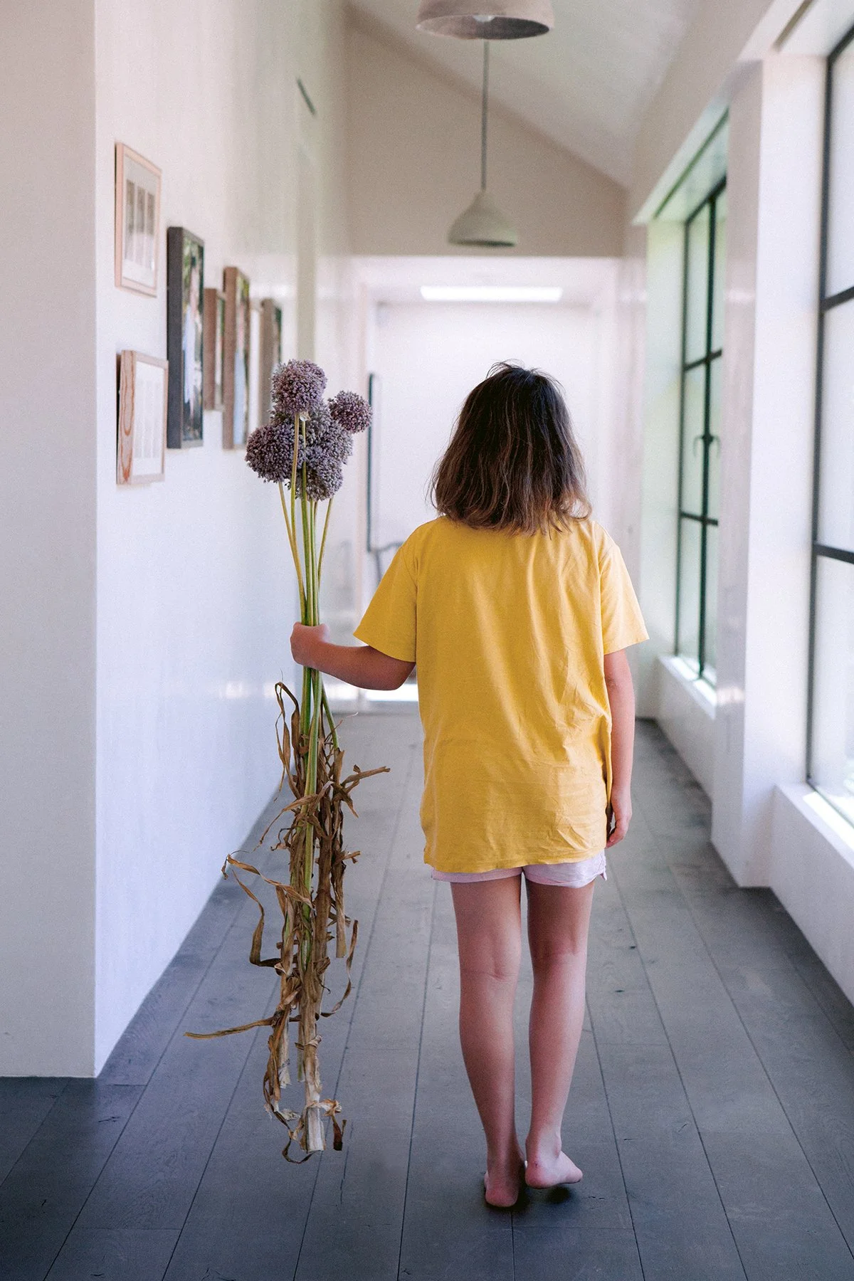 A girl with short hair wearing a yellow t-shirt and pink shorts walking barefoot away from the camera, down a brightly lit hallway holding a bunch of large purple allium flowers.