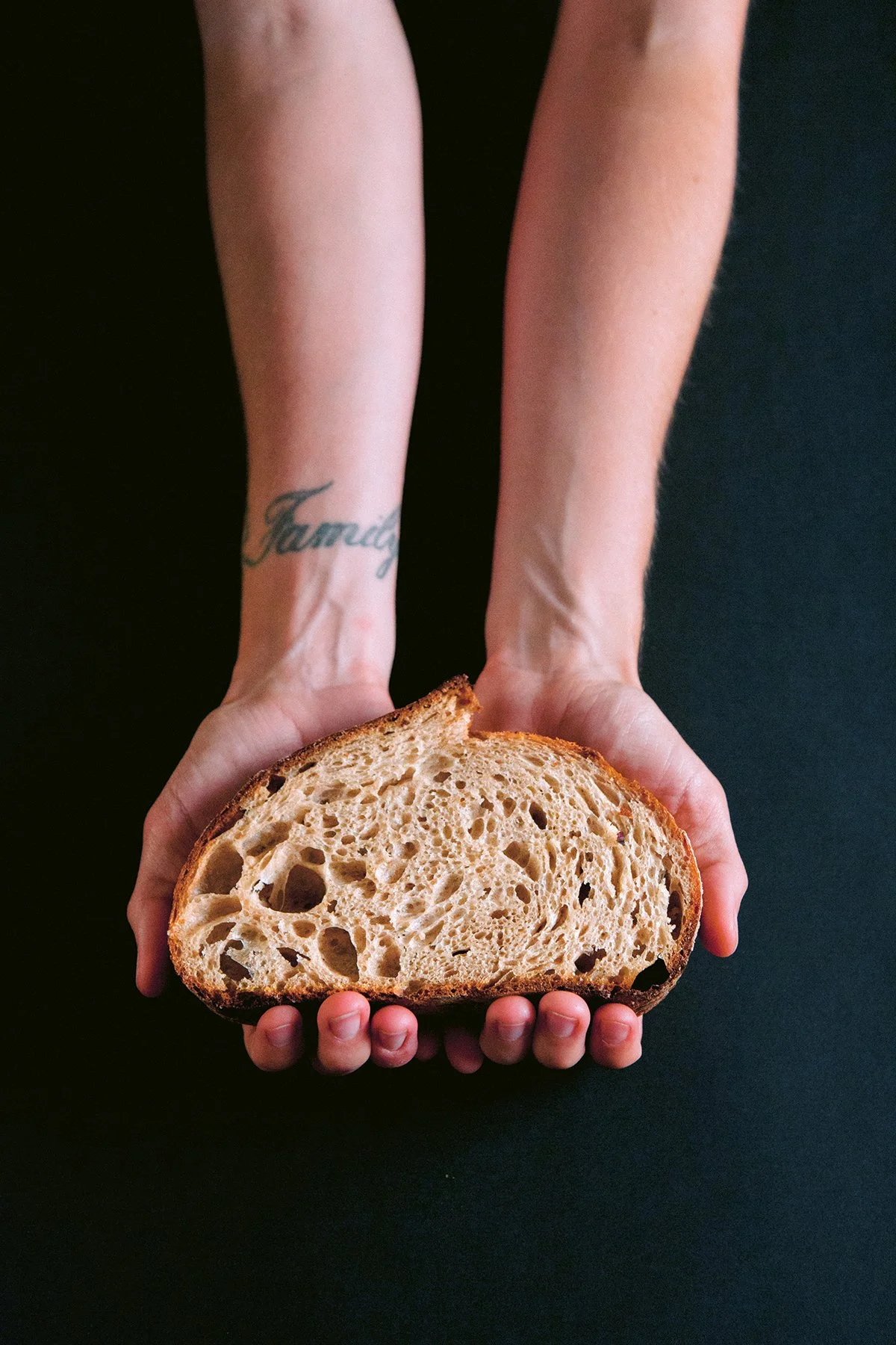 Woman's hands holding a large slice of artisan sourdough bread with a dark background. A tattoo that says "family" is on her wrist.