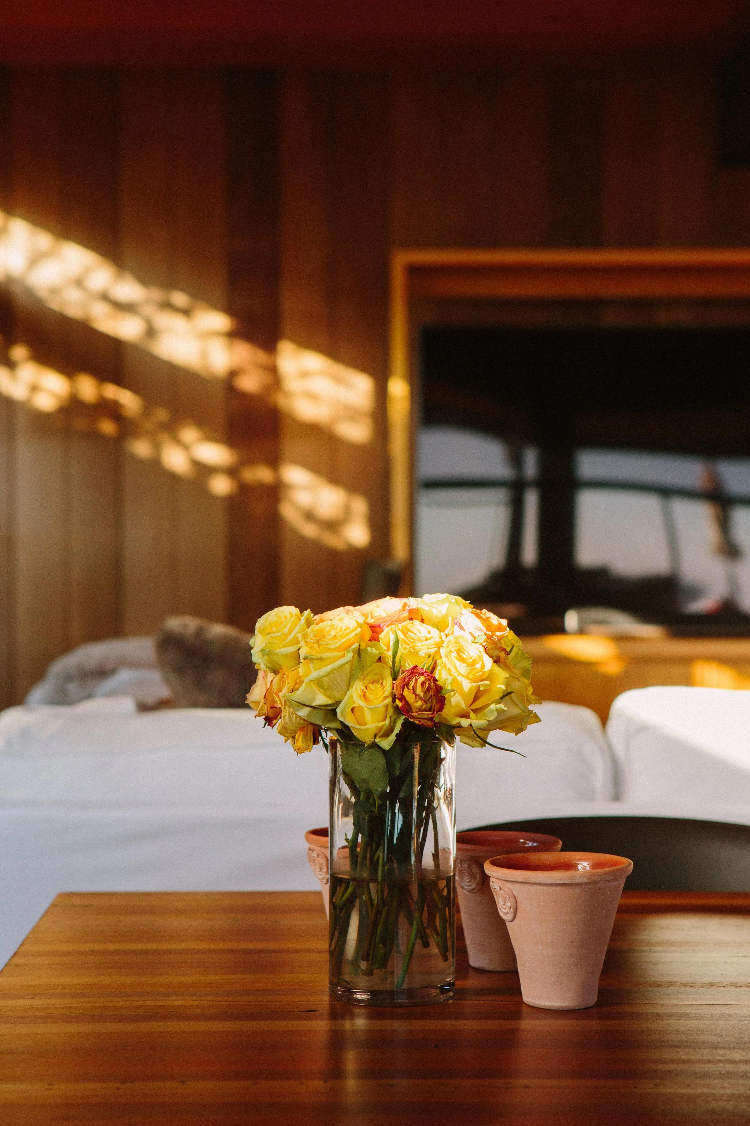 A bouquet of yellow and orange roses in a glass vase on a wooden table with two terracotta pots nearby, against a background of a dimly lit room with wooden paneling and a window with reflections at a beachfront private home in Malibu.