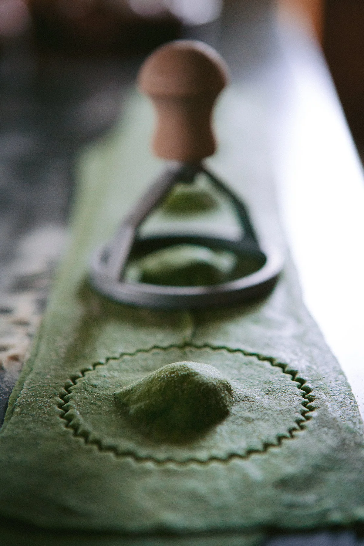 Close-up of a metal ravioli stamp pressing into fresh nettle pasta dough, creating a circular shape with an embossed edge.