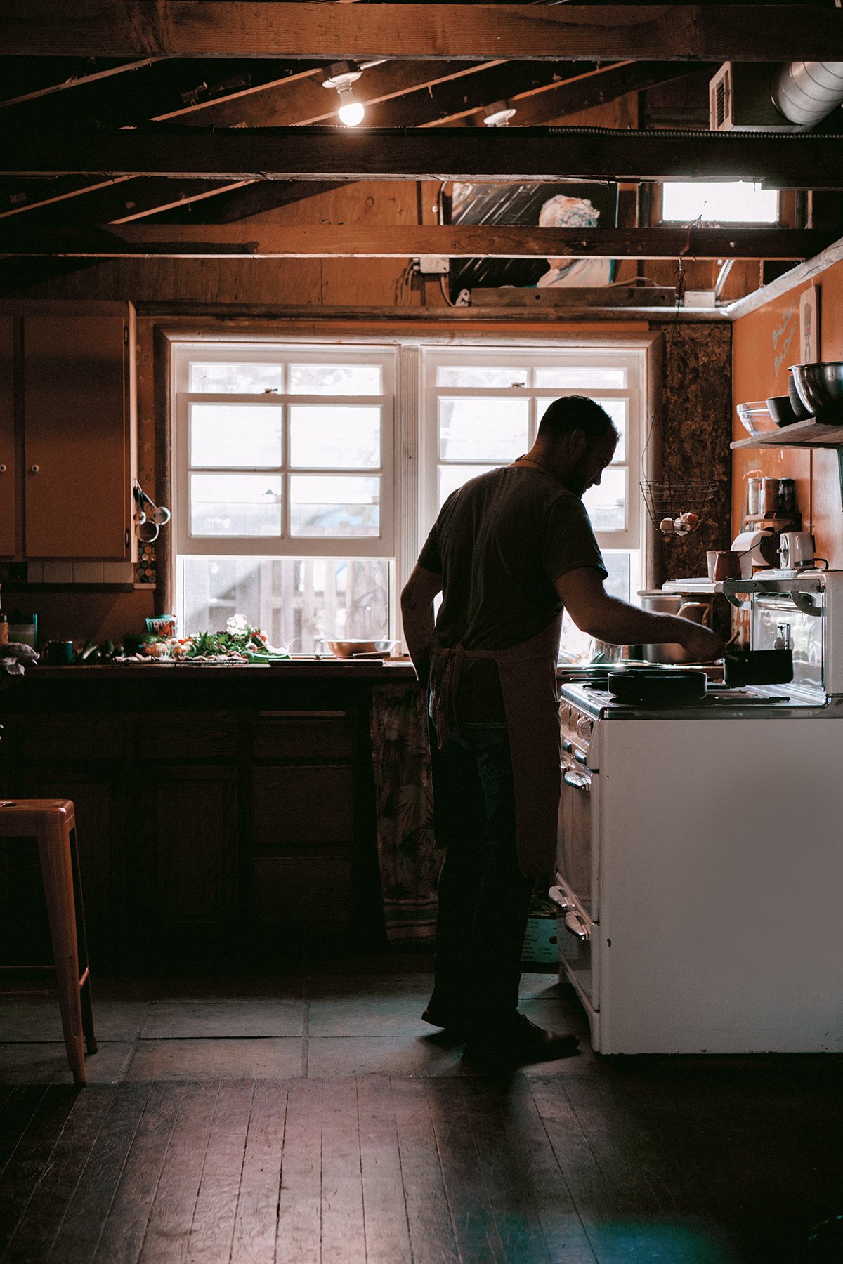 A chef cooking in a rustic kitchen with wooden walls and floors, standing at the stove. There are two large windows behind them letting in natural light / backlight,  and various kitchen items / utensils are visible on shelves and countertops.