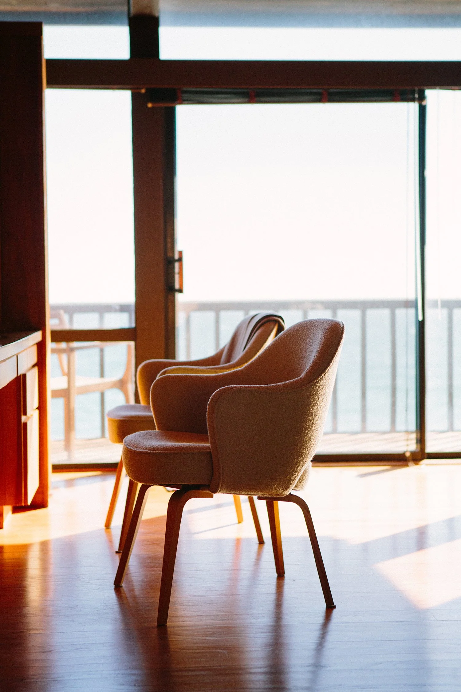 1970's Modern beachfront interior room with two beige upholstered chairs with wooden legs arranged around a low, wooden table. Large glass sliding doors in the background let in natural light and show a view of a balcony with a railing in Malibu.