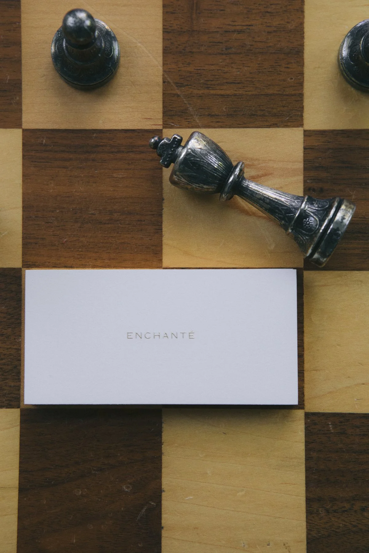 A chessboard with a fallen king piece, two pawns, and a card with the word 'ENCHANTÉ' on it.