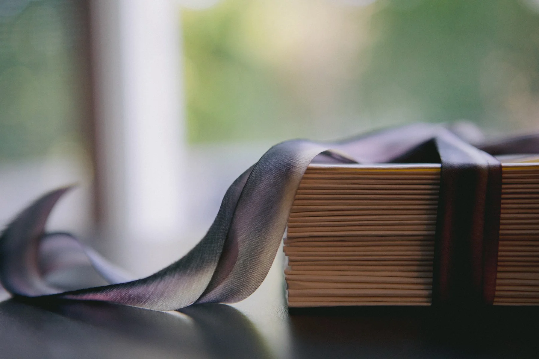 A stack of books with a dark ribbon bookmark on top, next to a dark satin ribbon, with a blurred window in the background.