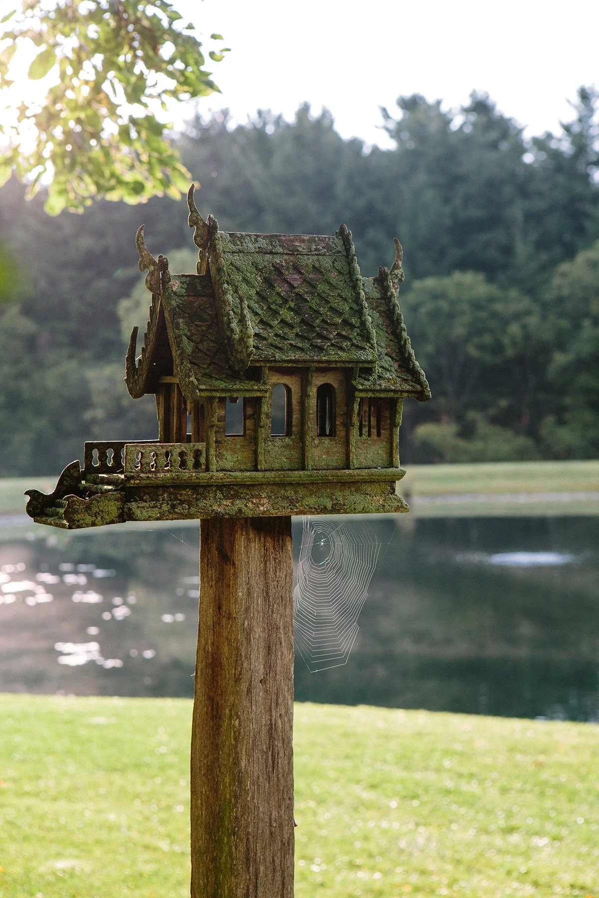 An old, moss-covered wooden Thai spirit house or house with intricate roof details, mounted on a wooden post near a body of water with grass and trees in the background, at a private estate in Lancaster Ohio.