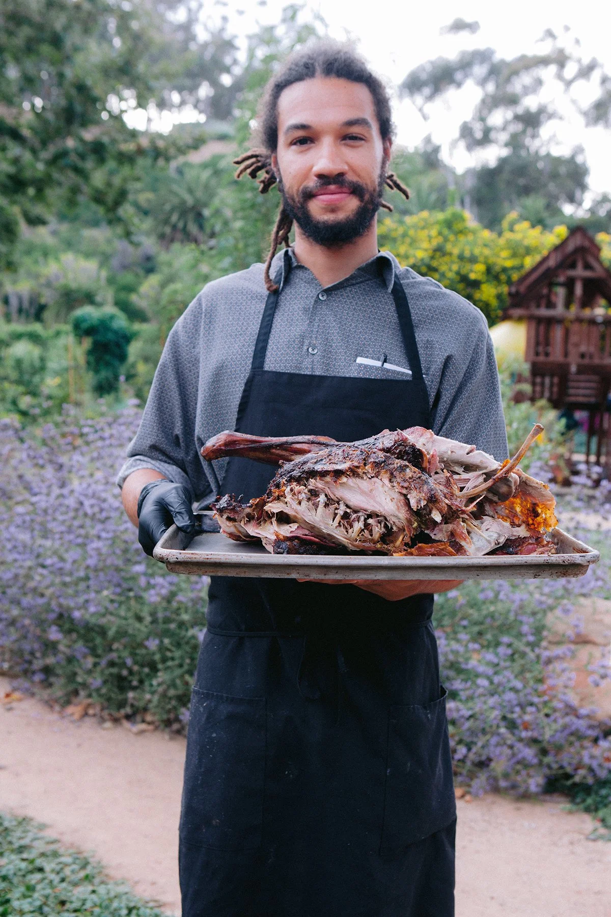 Man with dreadlocks and beard holding a tray of smoked meat outdoors at Plumcot Farm.